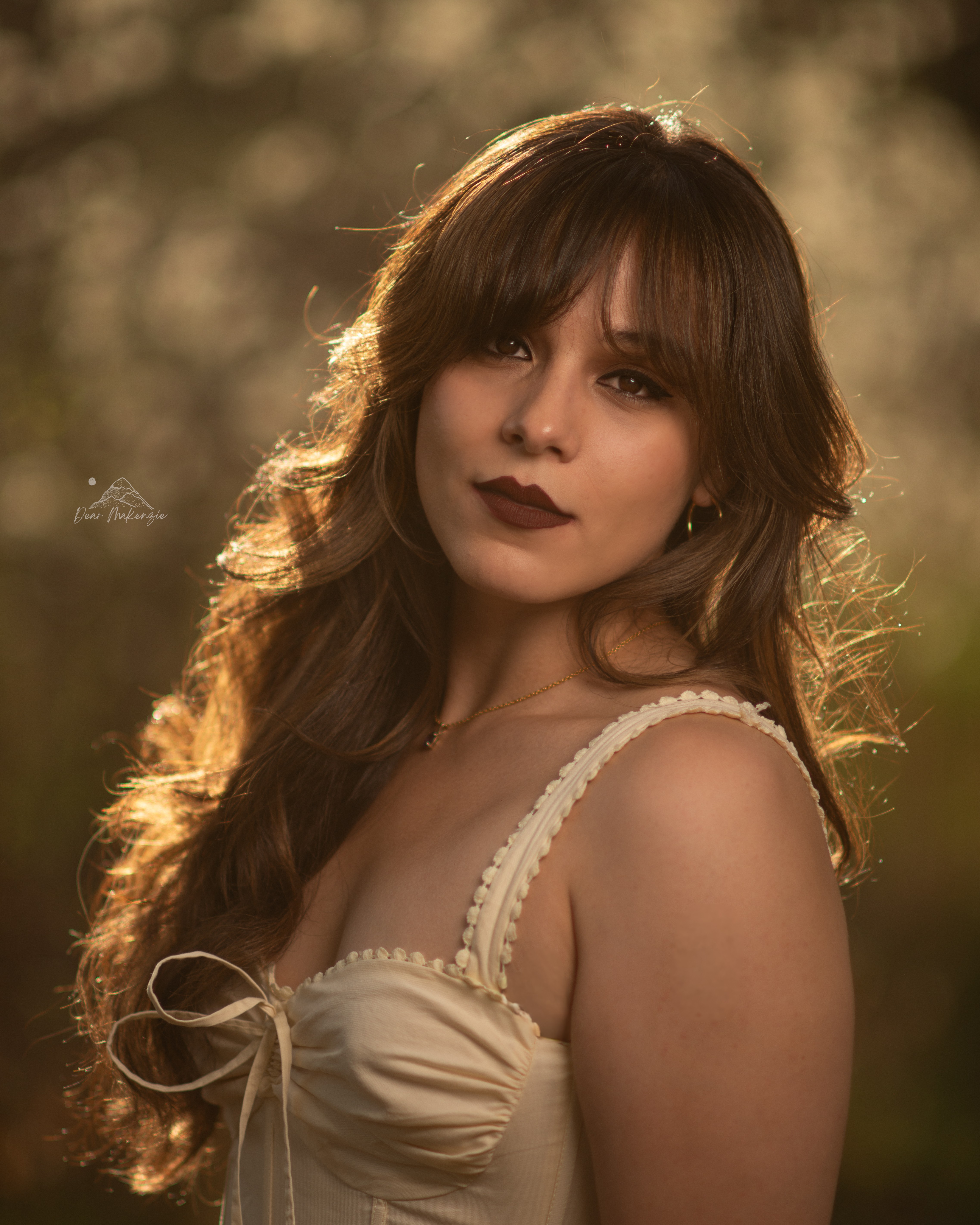 Young woman with long wavy hair and dark lipstick outdoors, sunlight behind her, wearing a cream-colored top with straps.