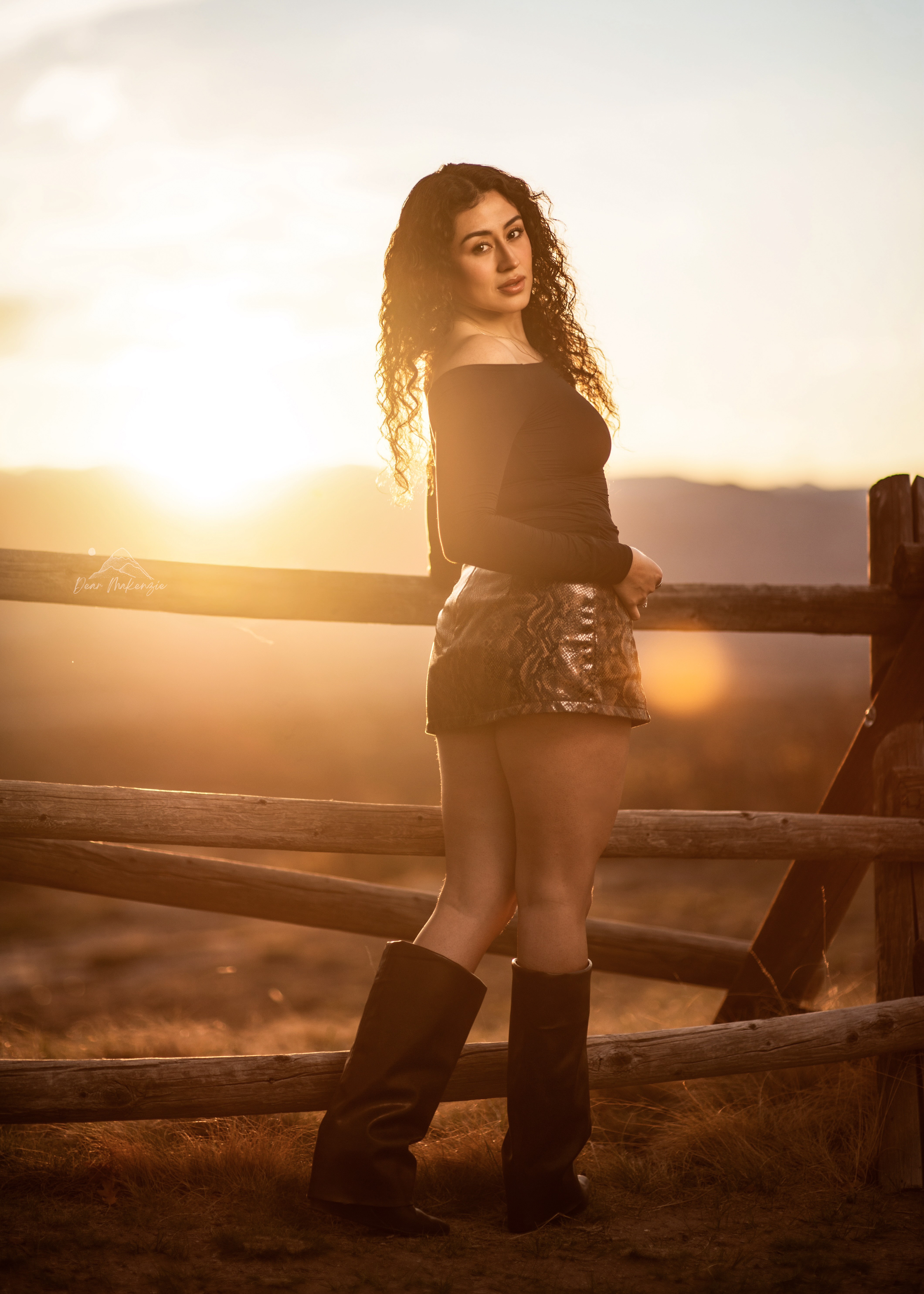 Young woman with curly hair standing outdoors near a wooden fence during sunset, wearing an off-shoulder top, shorts, and boots.