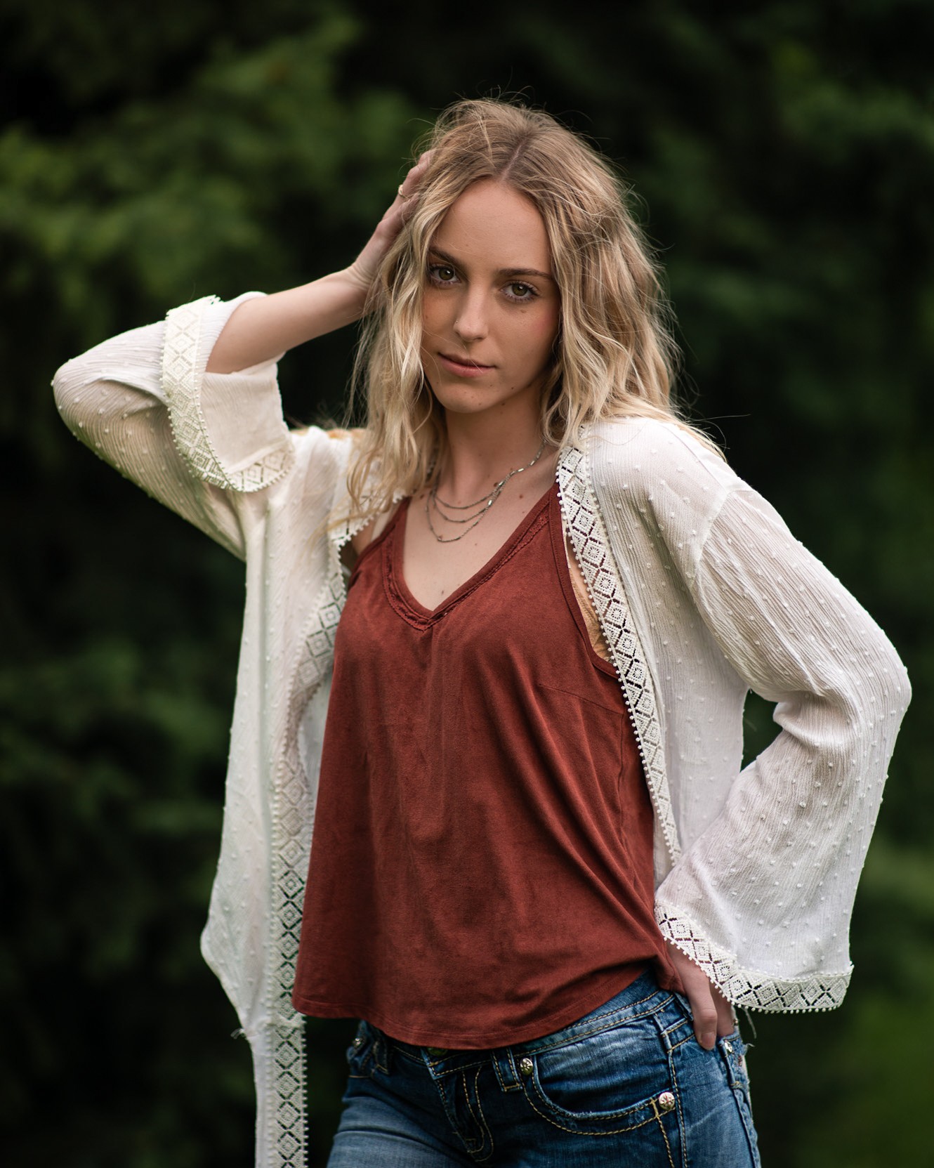 Young woman with wavy blonde hair outdoors, wearing a white cardigan over a rust-colored top and blue jeans.