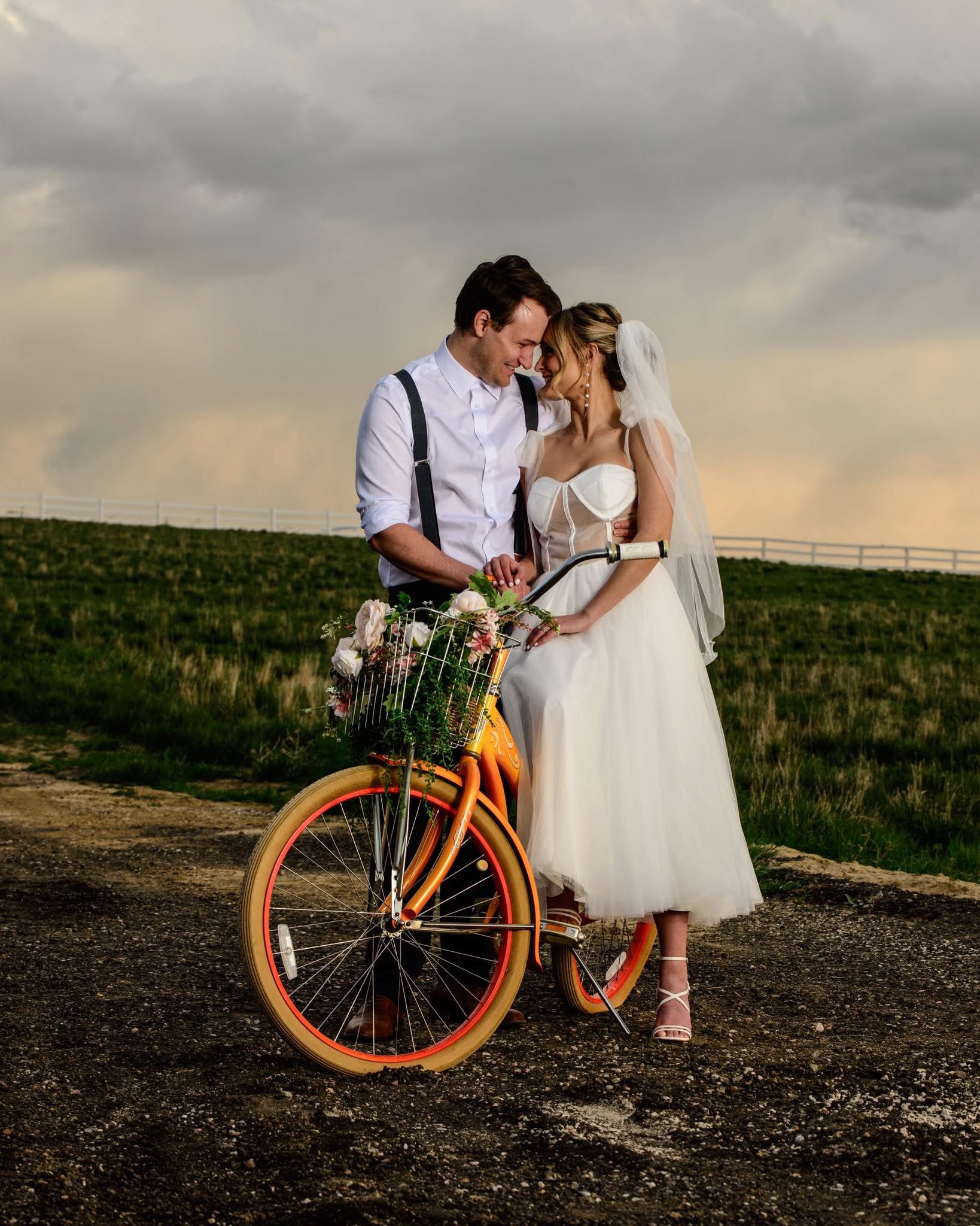 A bride and groom stand close together outdoors, with a bicycle and a field in the background, under cloudy sky.