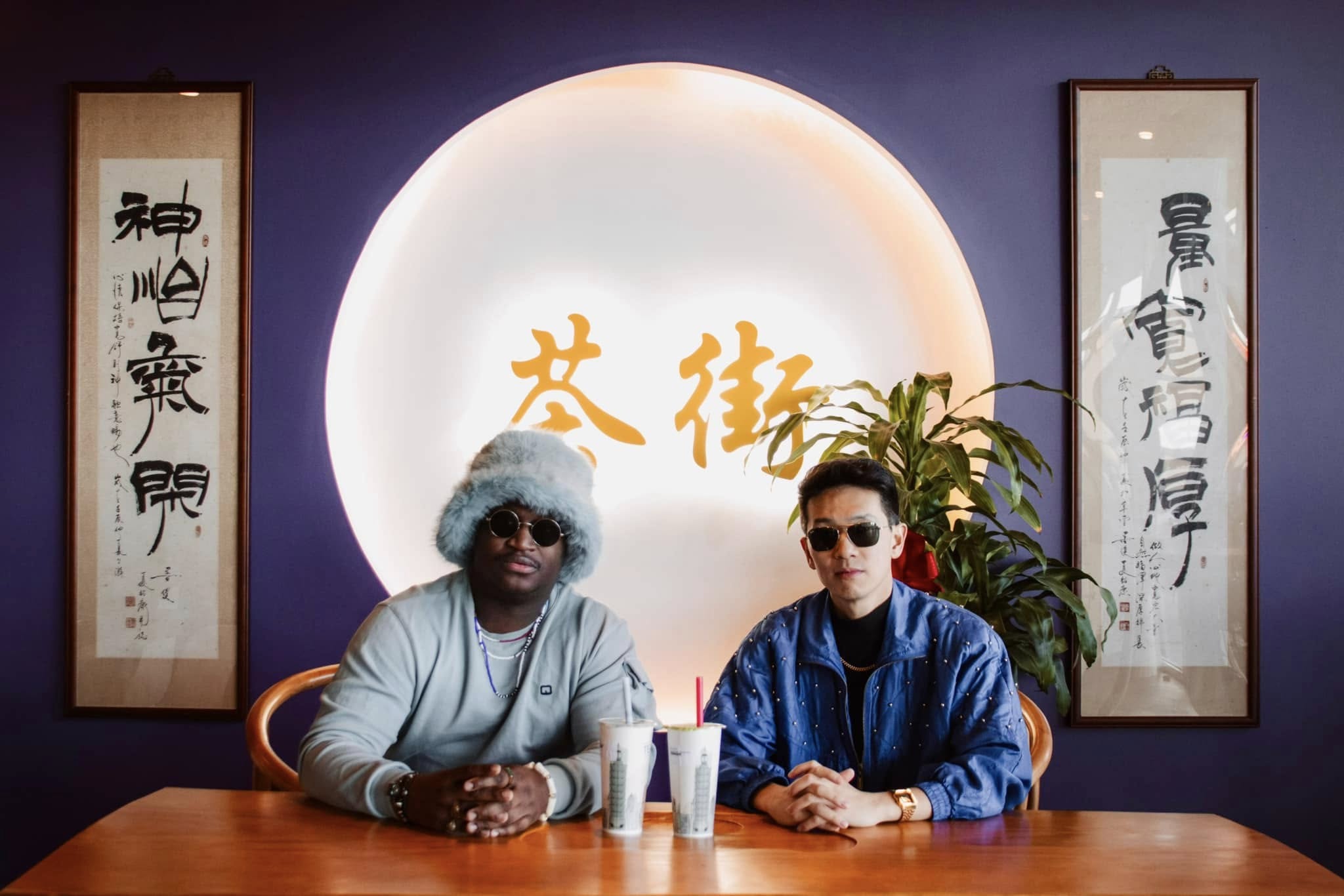 Two people sit at a table in front of a large illuminated circle with Chinese characters, flanked by calligraphy scrolls.