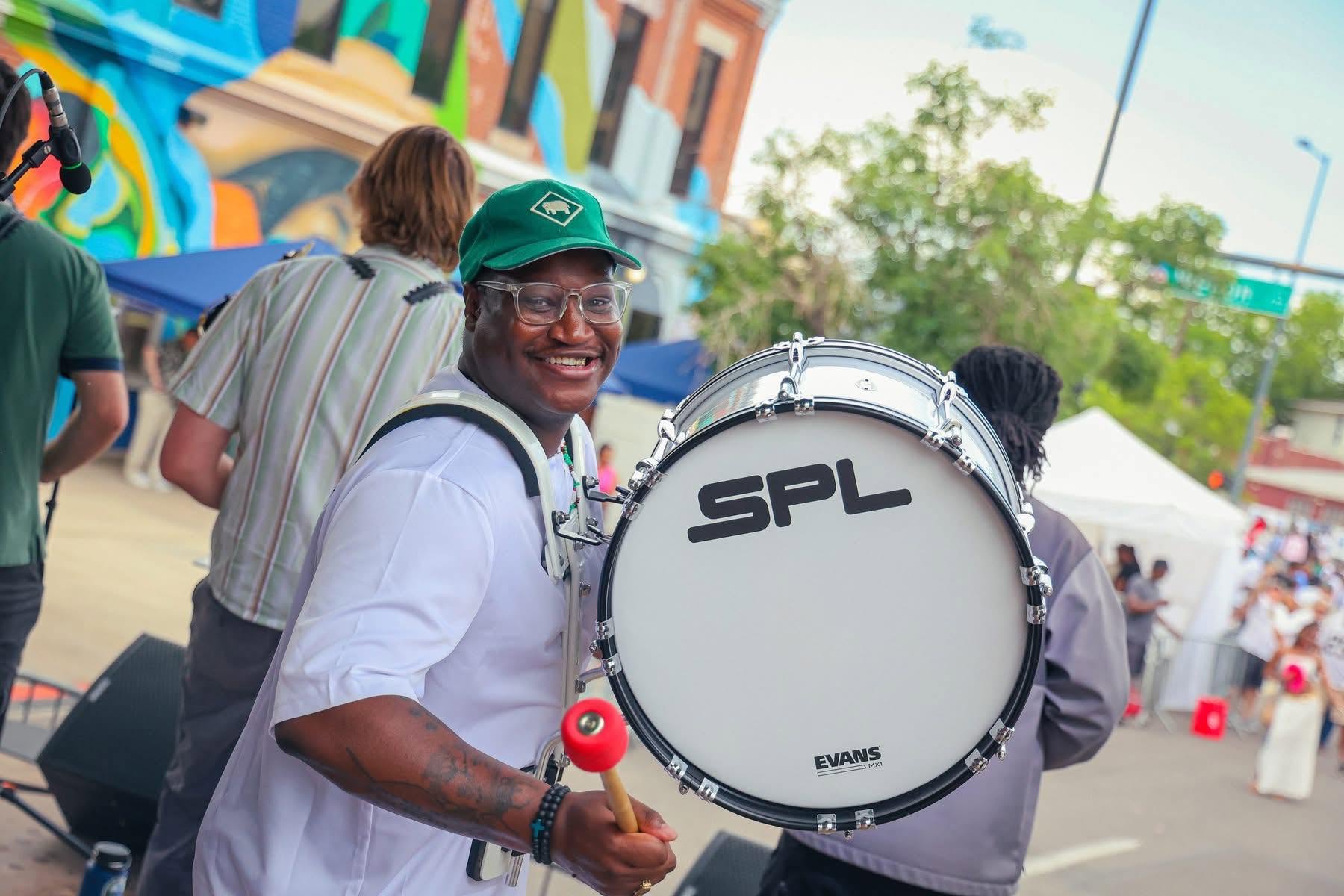Smiling man with glasses and a green cap holding a drumstick, standing outdoors with people and tents in background.