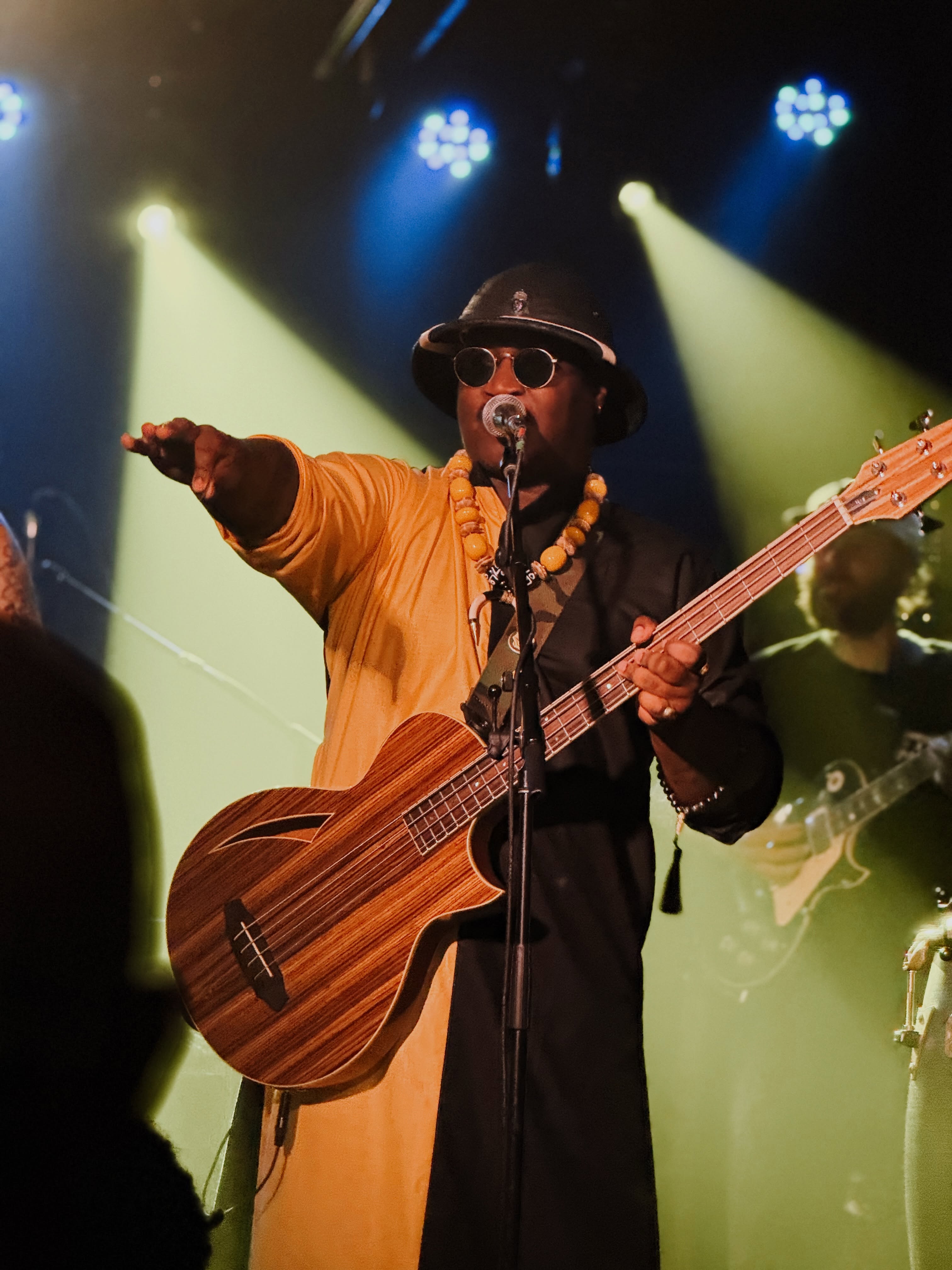 Musician on stage wearing sunglasses, hat, and yellow outfit, playing guitar and pointing, with stage lights behind.