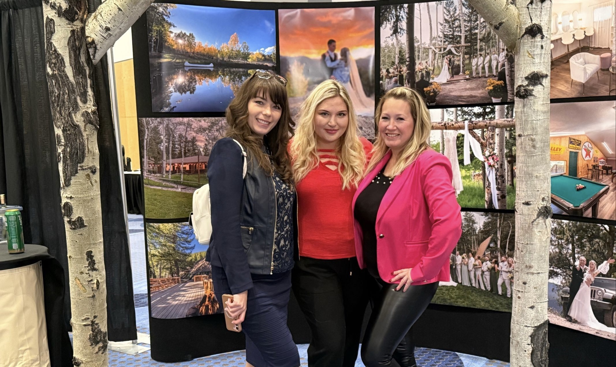 Three women standing together in front of a display of landscape photographs, smiling and posing for the camera.