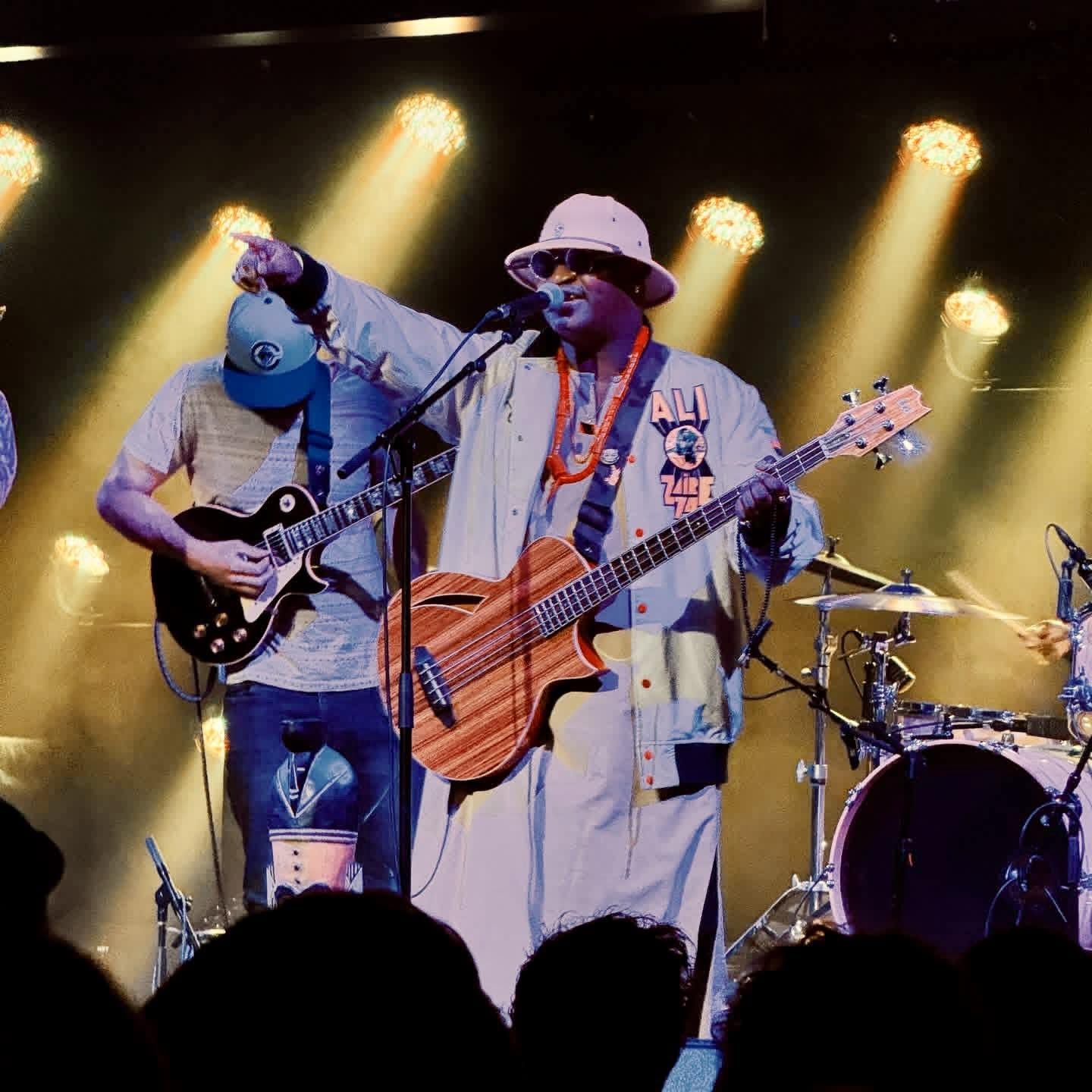 Performer with a guitar, wearing a hat and sunglasses, on stage with band members and drums, stage lights shining.