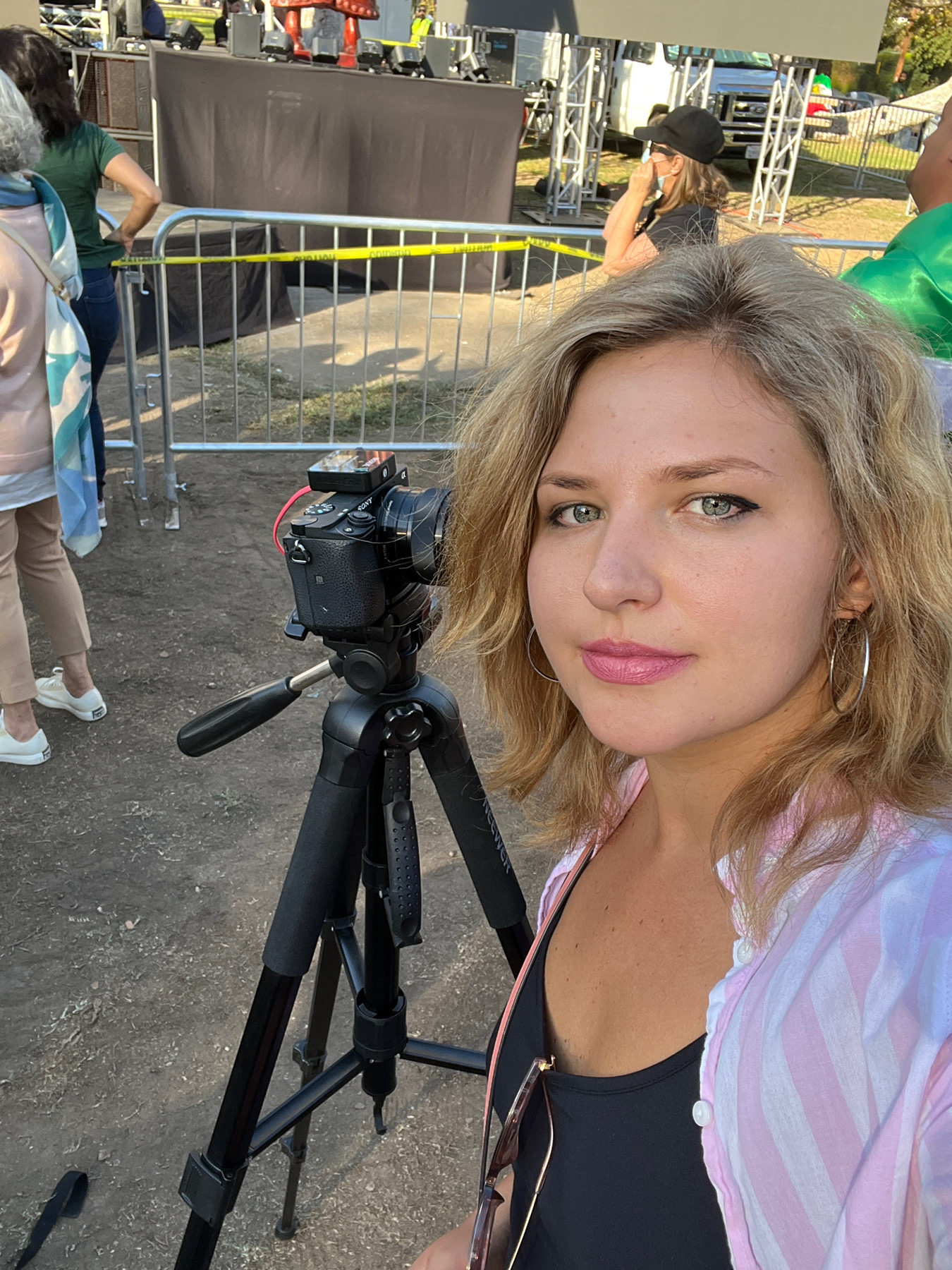 Young woman with blonde hair and pink lipstick takes a selfie at an outdoor event, with a camera on a tripod behind her.
