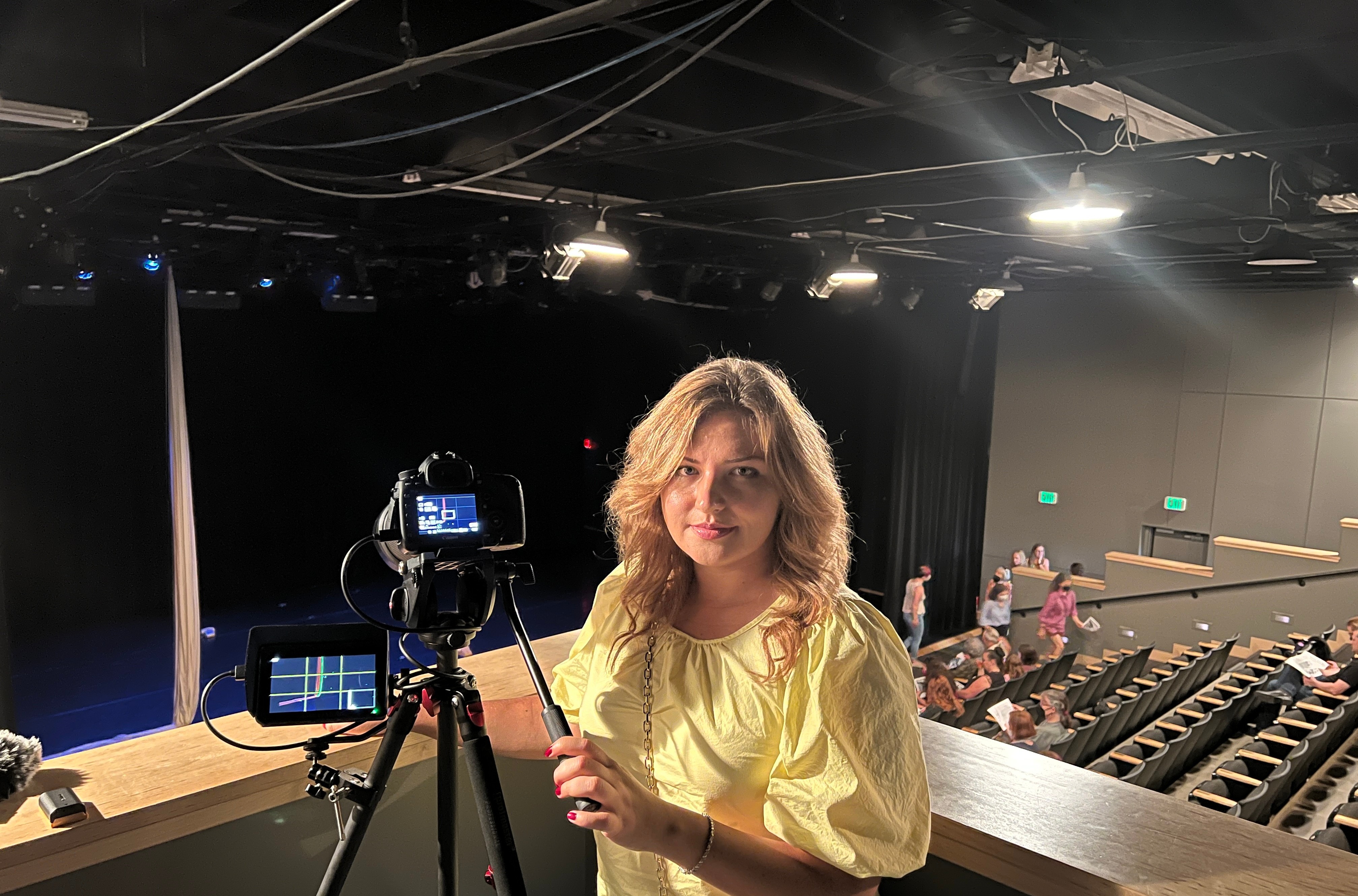 Woman with curly hair in a yellow top standing in a theater or studio with camera equipment, audience seats, and stage in background.