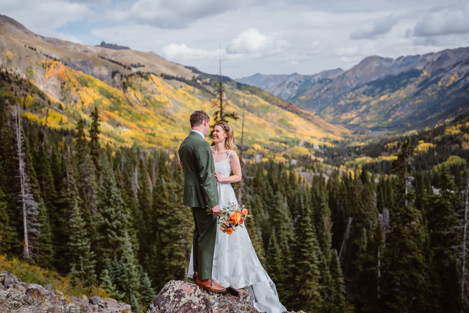 A couple in wedding attire stands on a mountain overlook with a forest and colorful mountains in the background.