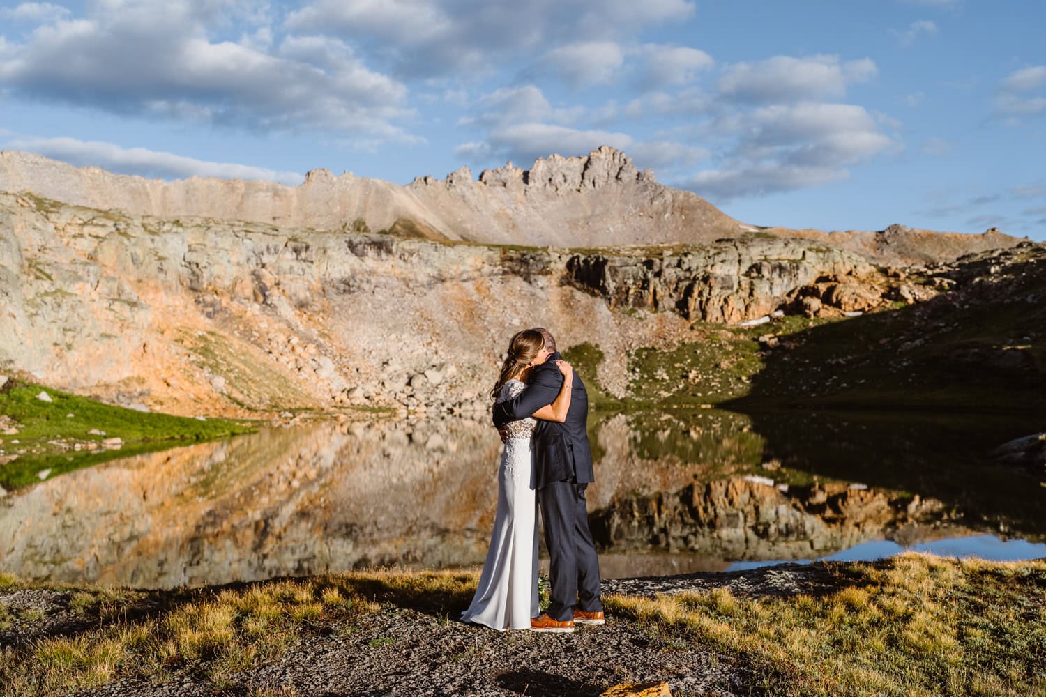 Two people embrace near a lake with mountains and a partly cloudy sky in the background.
