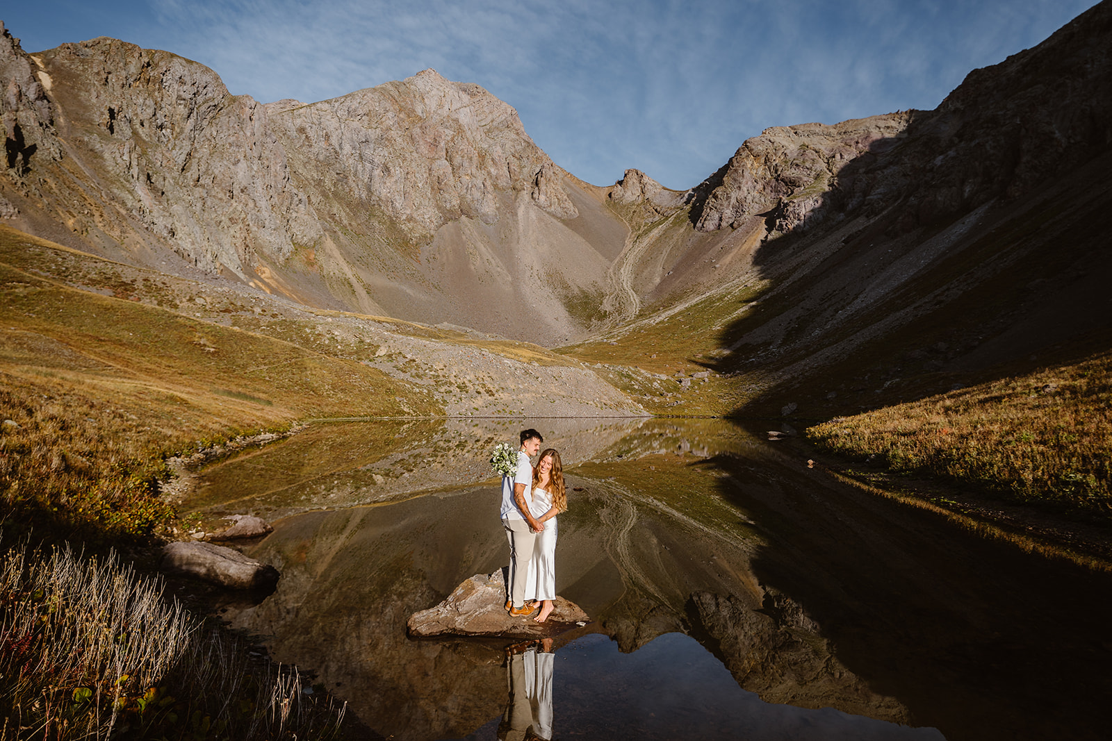 A couple stands on a rock in a mountain lake, surrounded by mountains and a blue sky, with their reflection in the water.