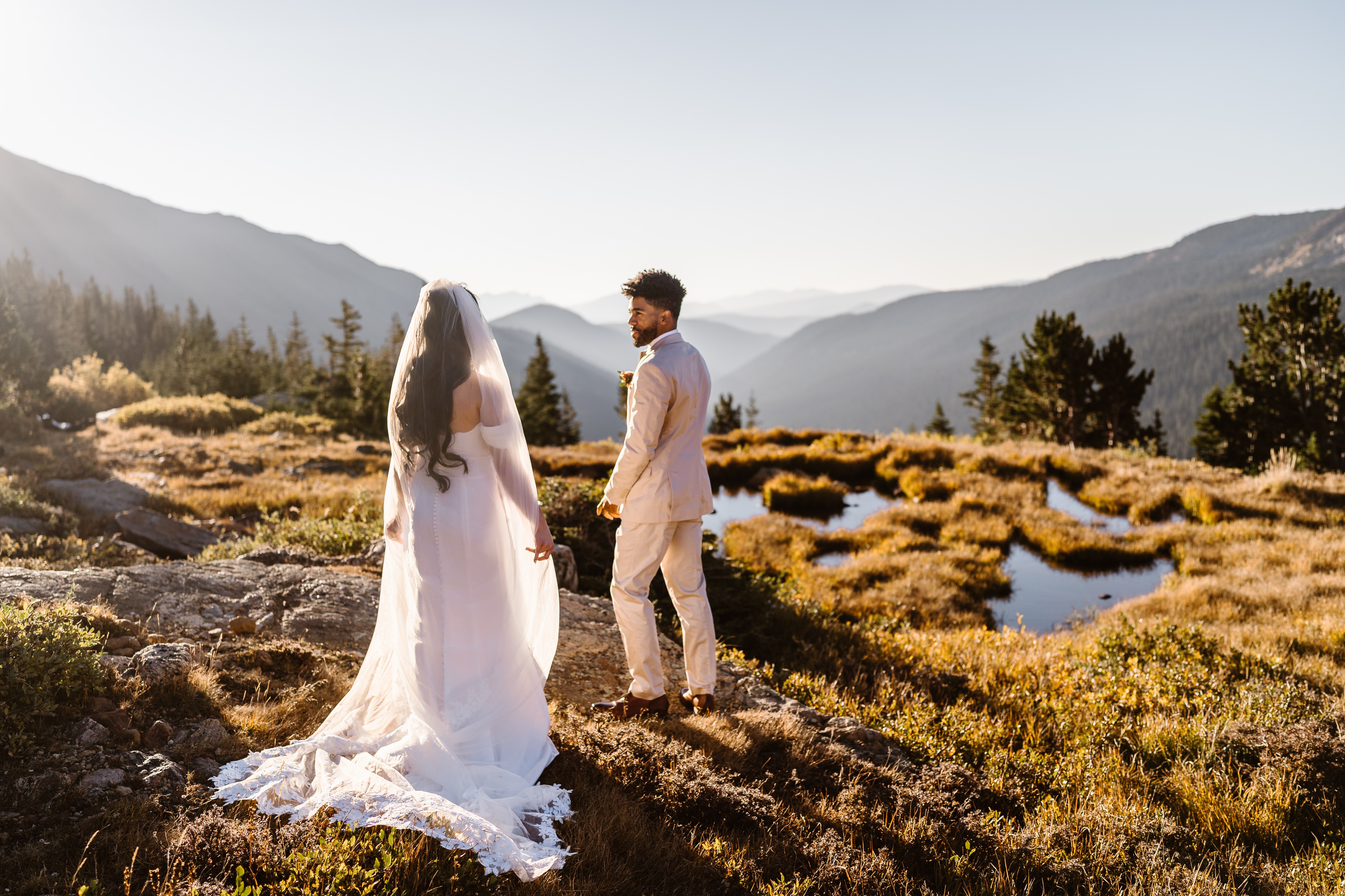 Bride and groom holding hands outdoors with mountains and trees in the background, sunlight illuminating the scene.