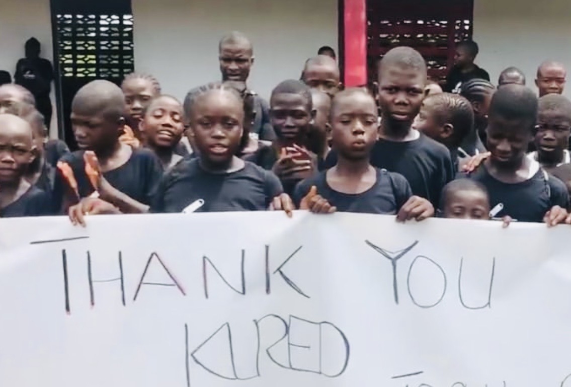 Group of children holding a sign that says 'THANK YOU KARED' outdoors.