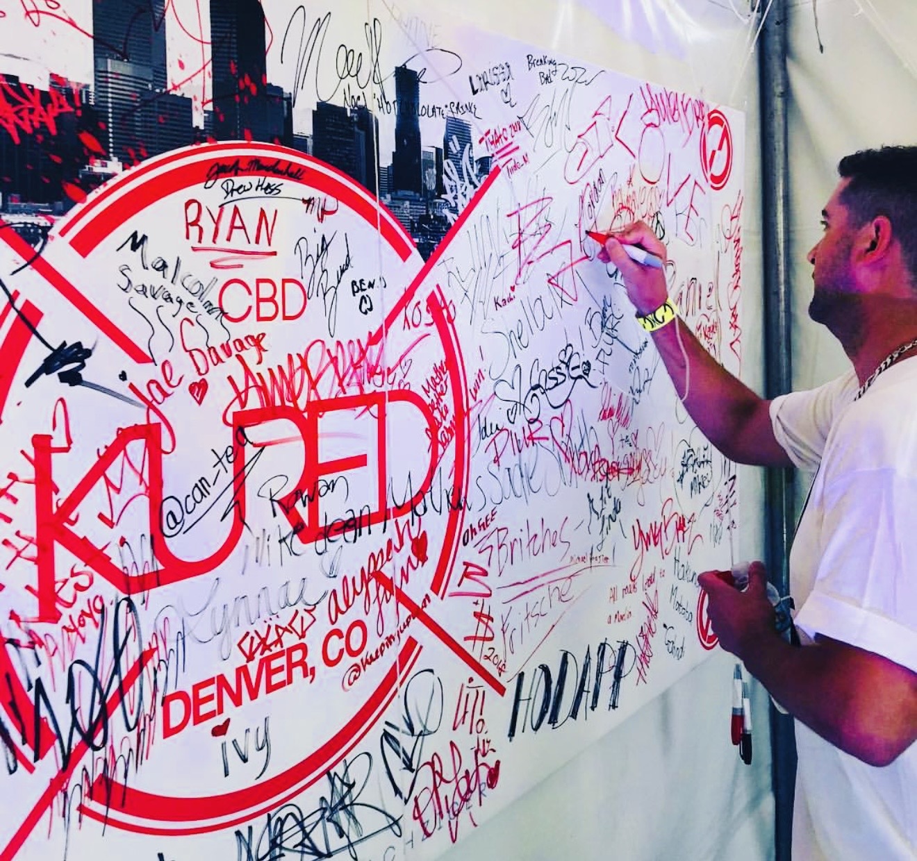 Person signing a large white banner with red and black text, covered in signatures and messages, outdoors.