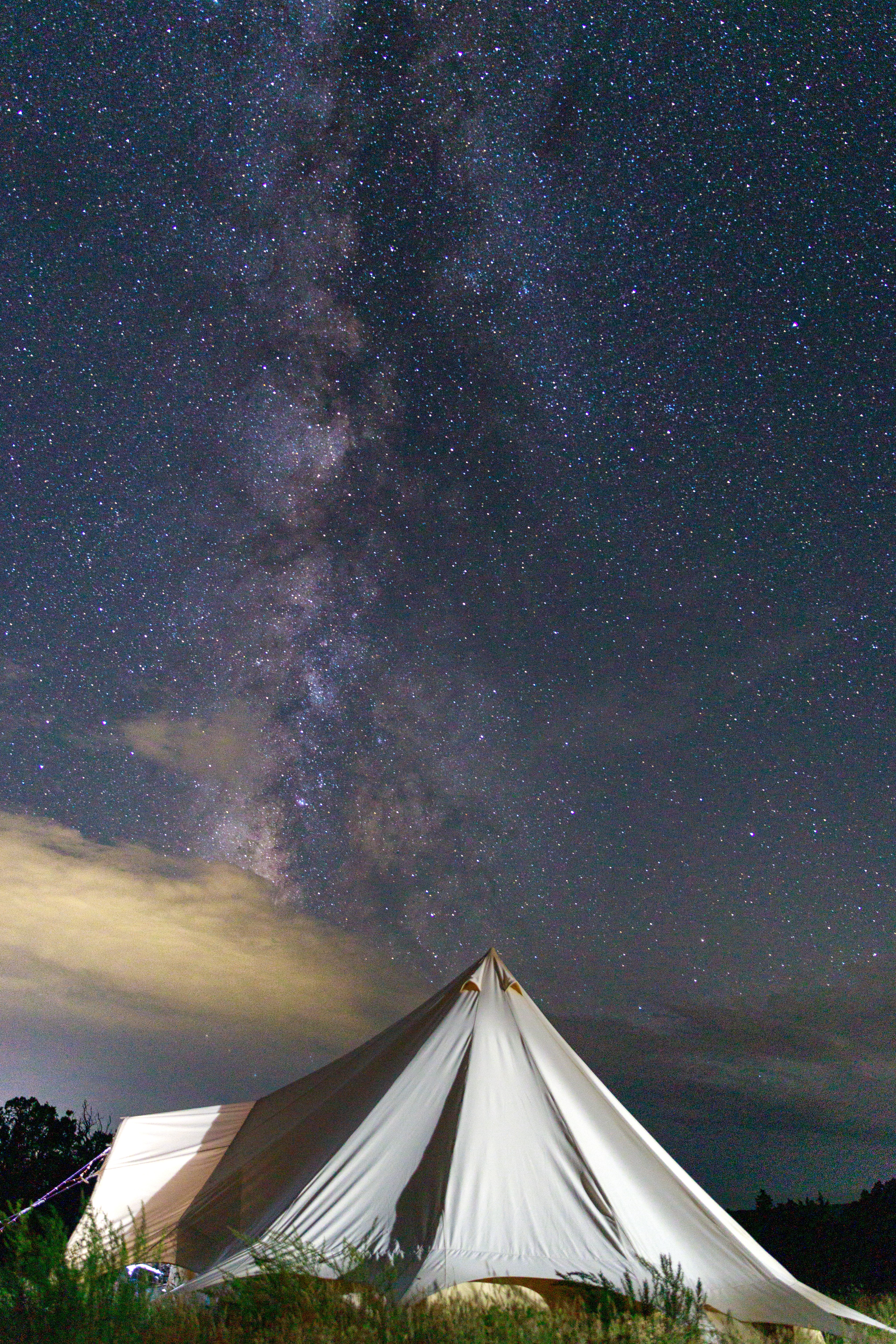 Night sky with stars and the Milky Way above a white tent near water, with clouds at the horizon.