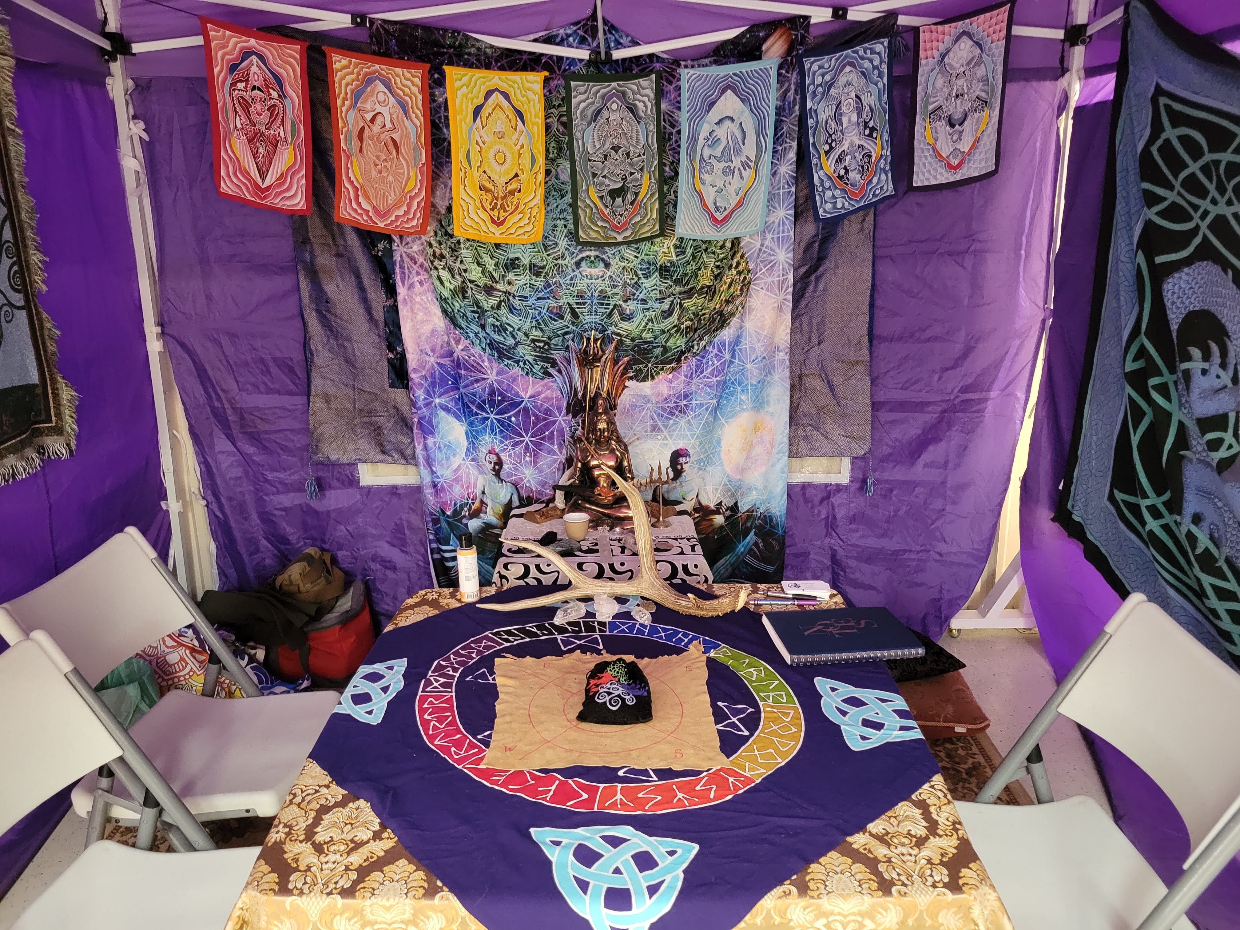 Table with a cloth featuring a circular design, surrounded by chairs, with colorful hanging banners and a tapestry in the background.
