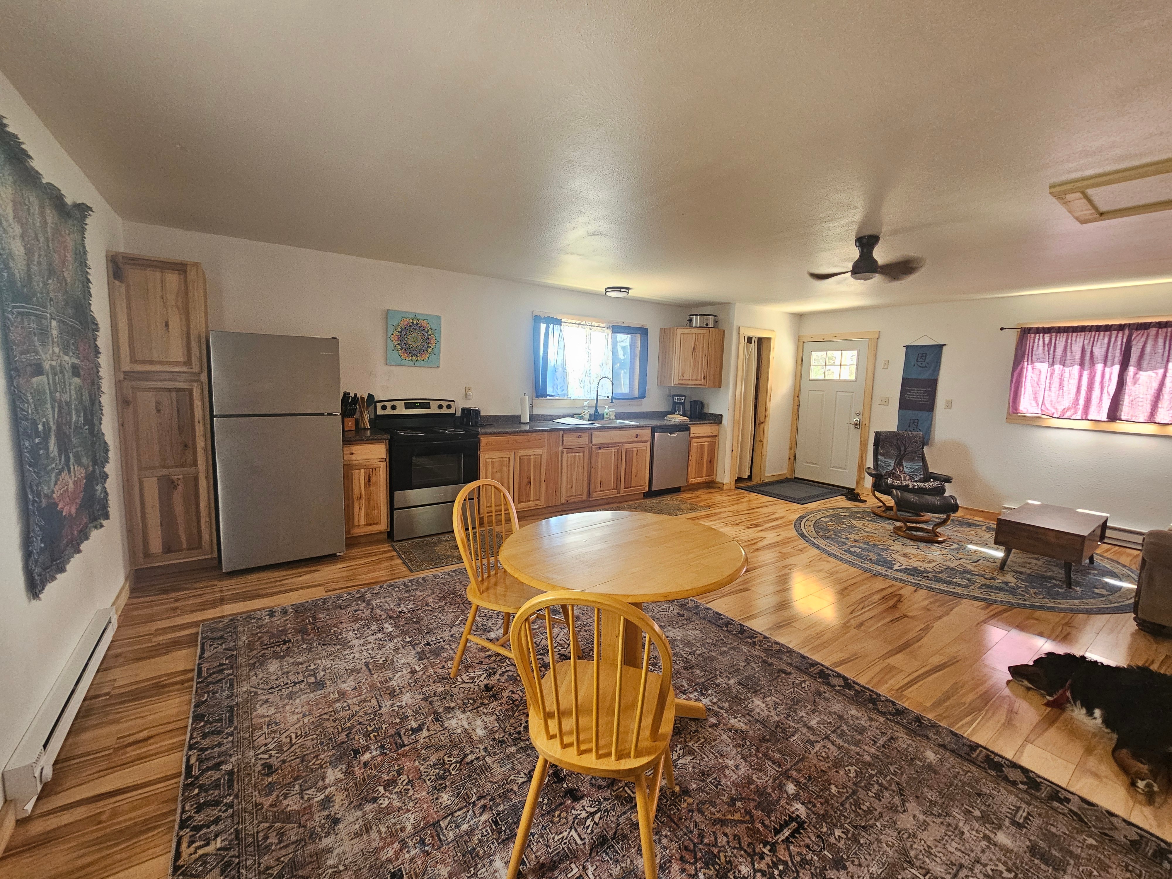 Living room with wooden cabinets, stainless steel refrigerator, round dining table with two yellow chairs, and a window with pink curtains.