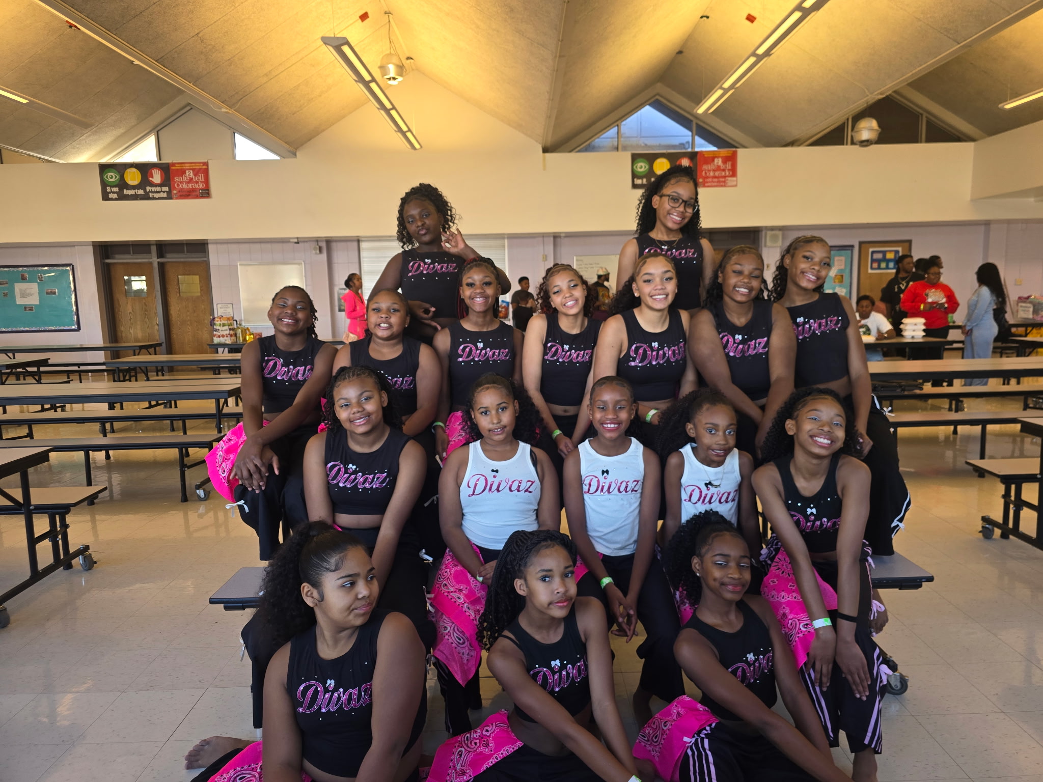 Group of young girls in dance costumes posing in a gymnasium with benches and a high ceiling.
