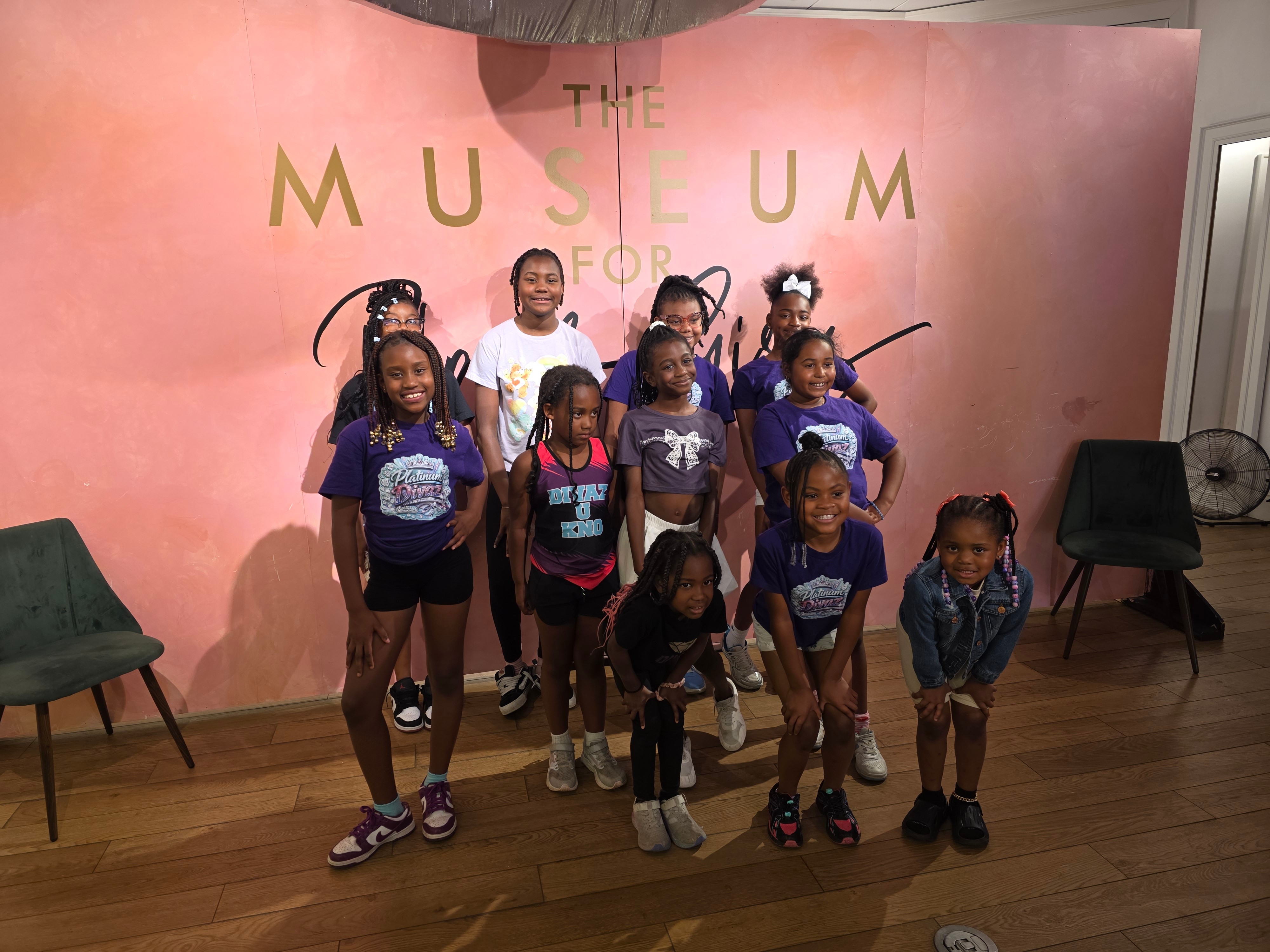 Group of children and an adult posing in front of a pink wall with gold letters, some children wearing purple shirts, some with backpacks, in a museum setting.