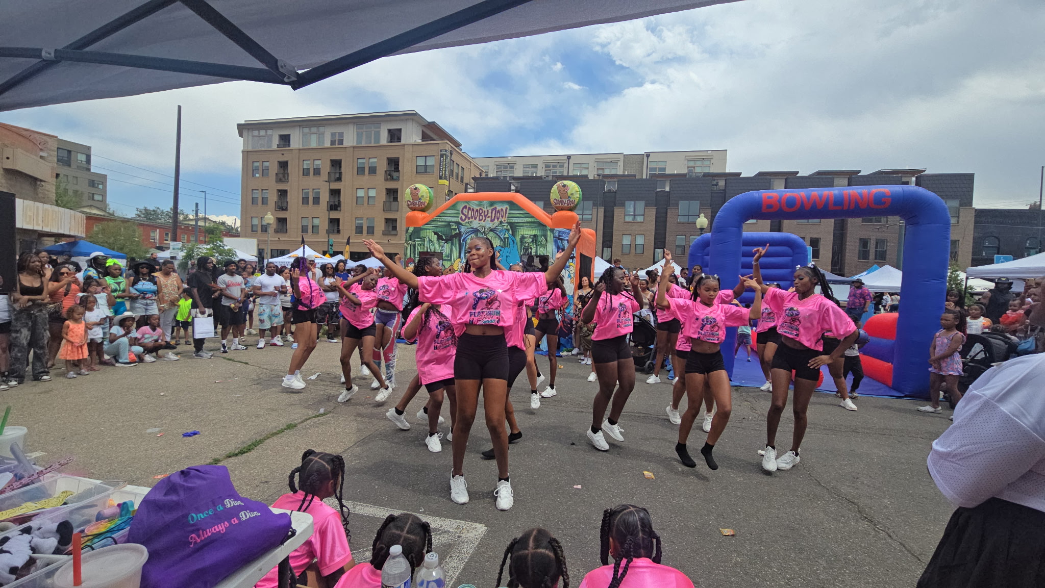 Group of people dancing outdoors with colorful inflatable structures in the background, crowd watching, and tents nearby.
