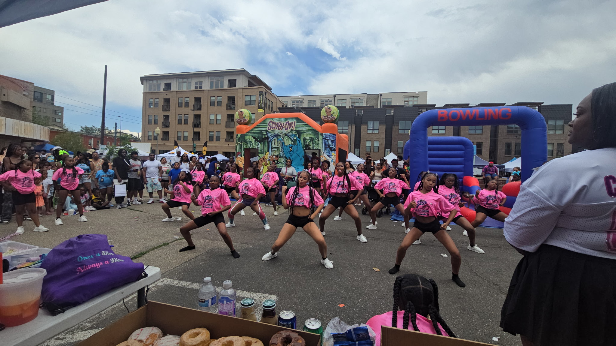 Group of people dancing outdoors with pink costumes, inflatable structures, and buildings in the background, under a partly cloudy sky.