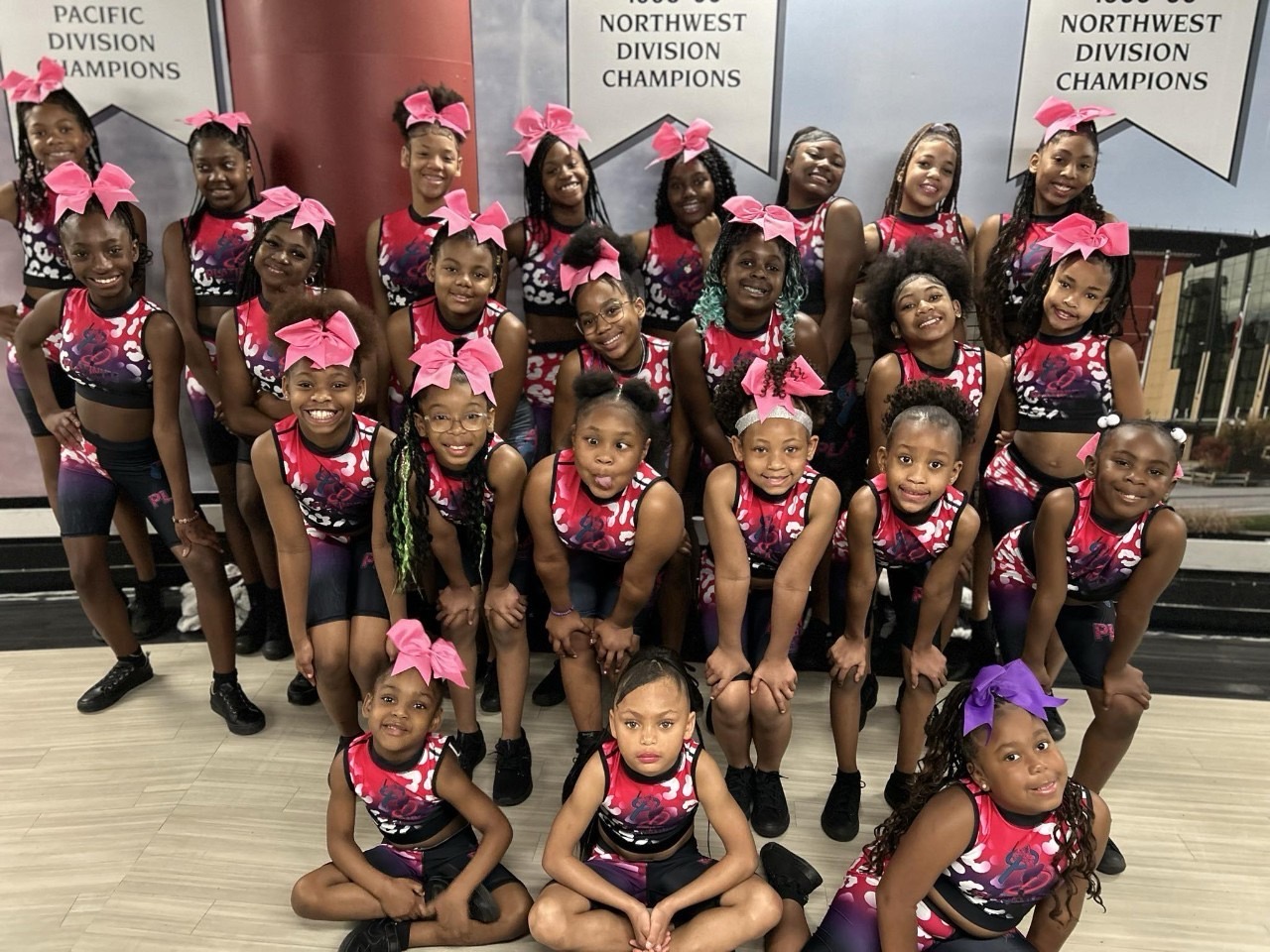 Group of young girls in cheerleading uniforms with pink bows, posing indoors with banners in the background.