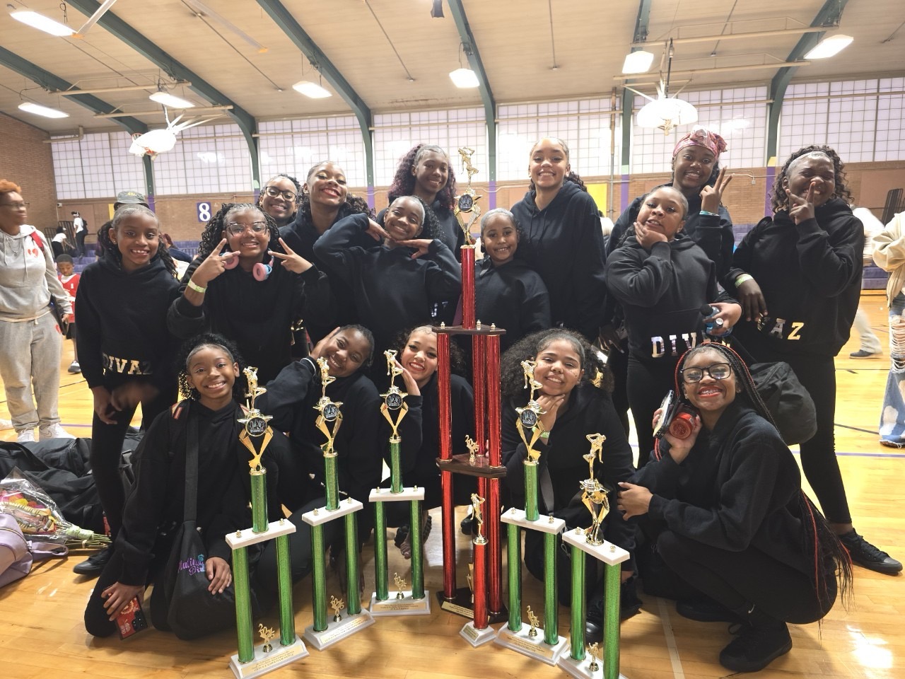 Group of young girls and women in black sports uniforms posing with trophies and a large red candle in a gymnasium.
