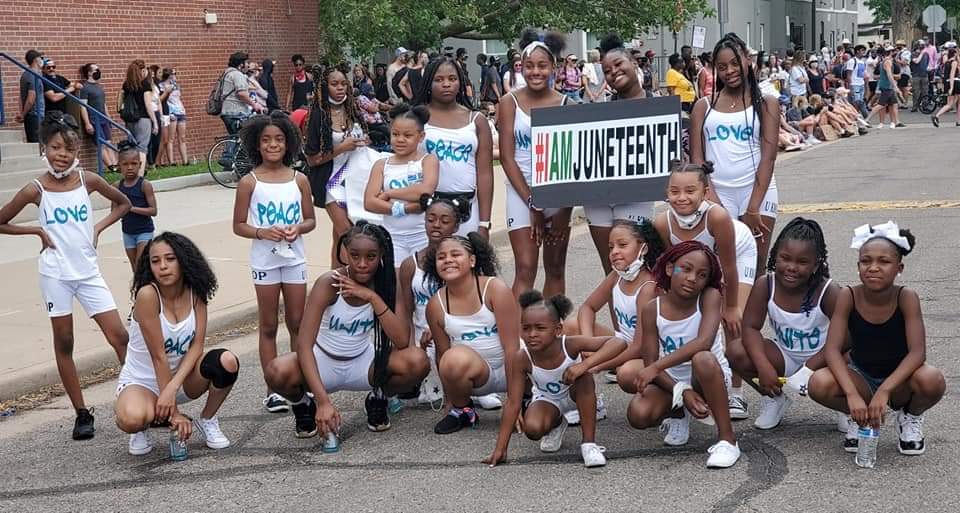 Group of young girls participating in a parade holding a banner, some wearing matching shirts, on a city street.