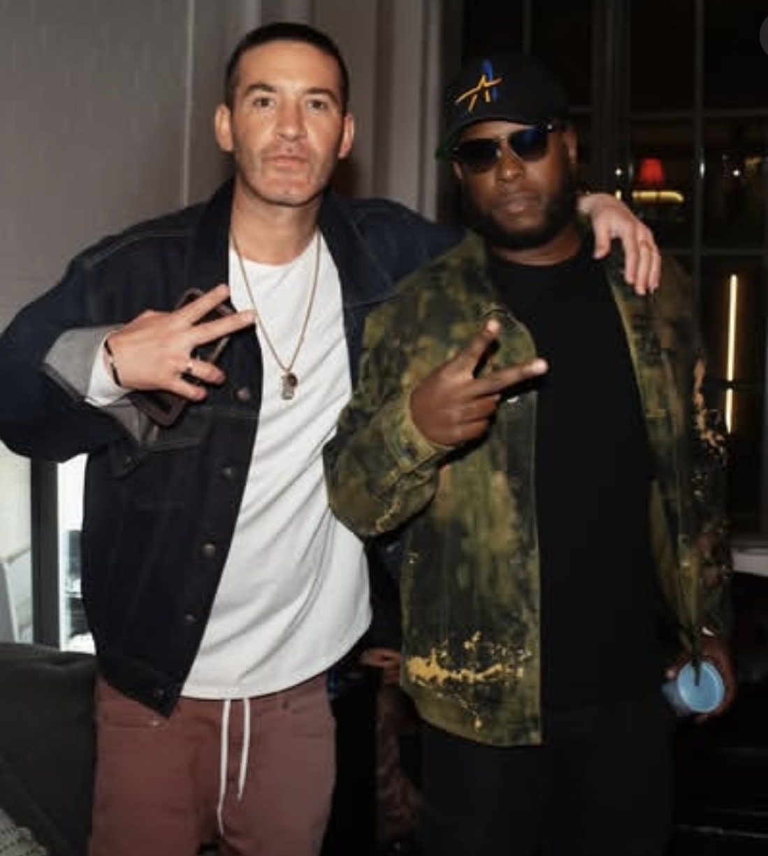Two men standing indoors, one with arm around the other, both making peace signs, wearing casual clothing and accessories.