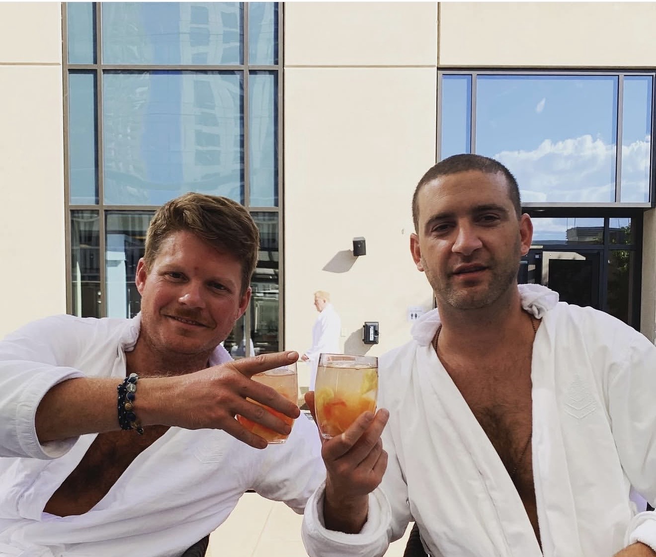 Two men in white shirts holding drinks outdoors with a modern building and blue sky in the background.