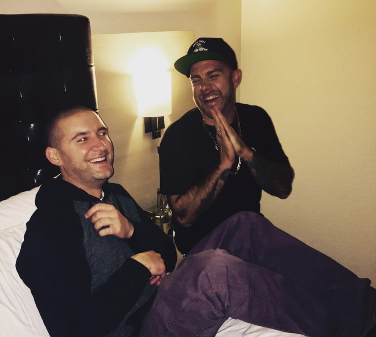 Two men sitting on a bed, smiling and laughing, in a warmly lit room with a lamp on the wall.