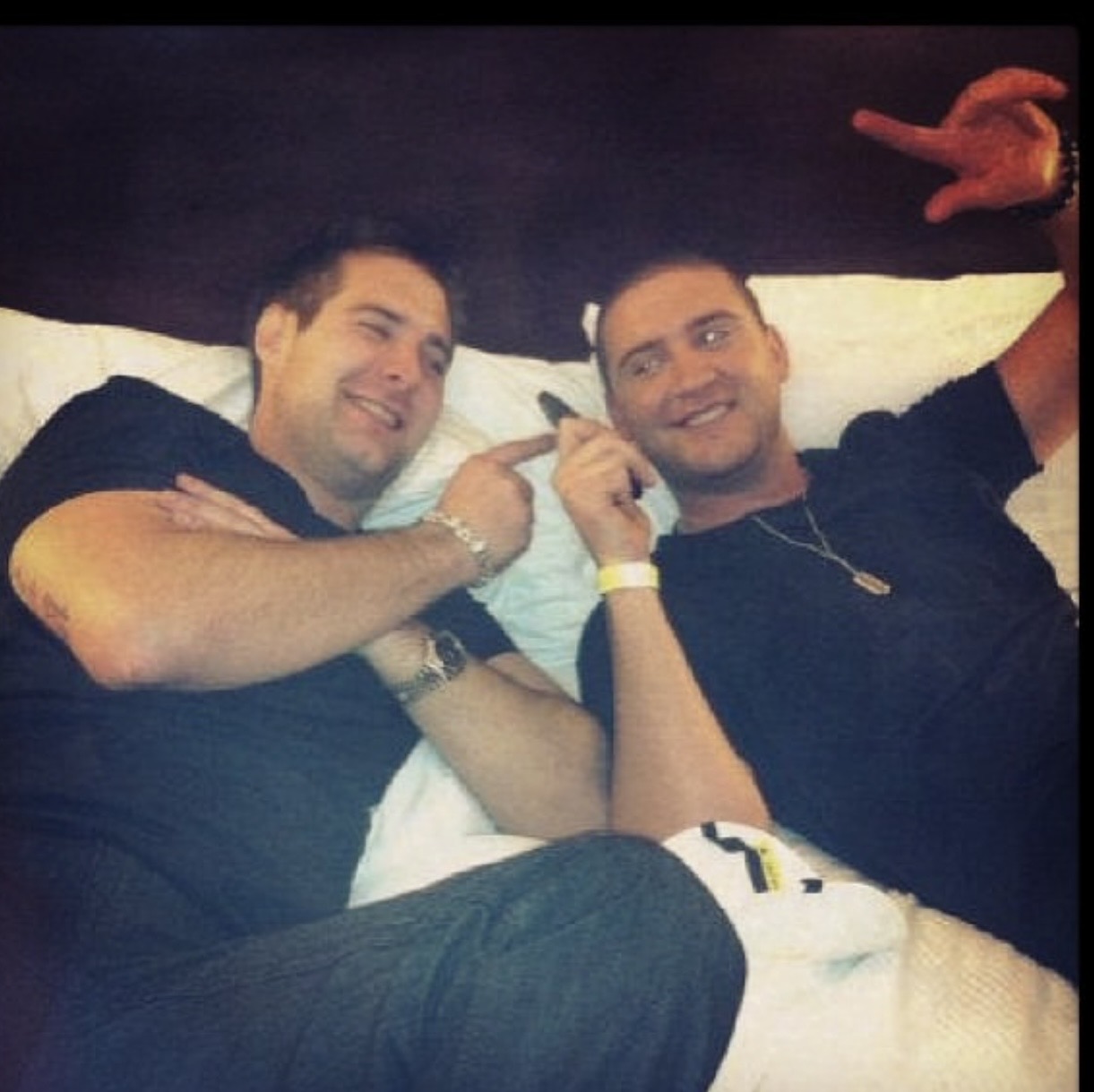 Two men lying on a bed, smiling and pointing at each other, with one making a peace sign.