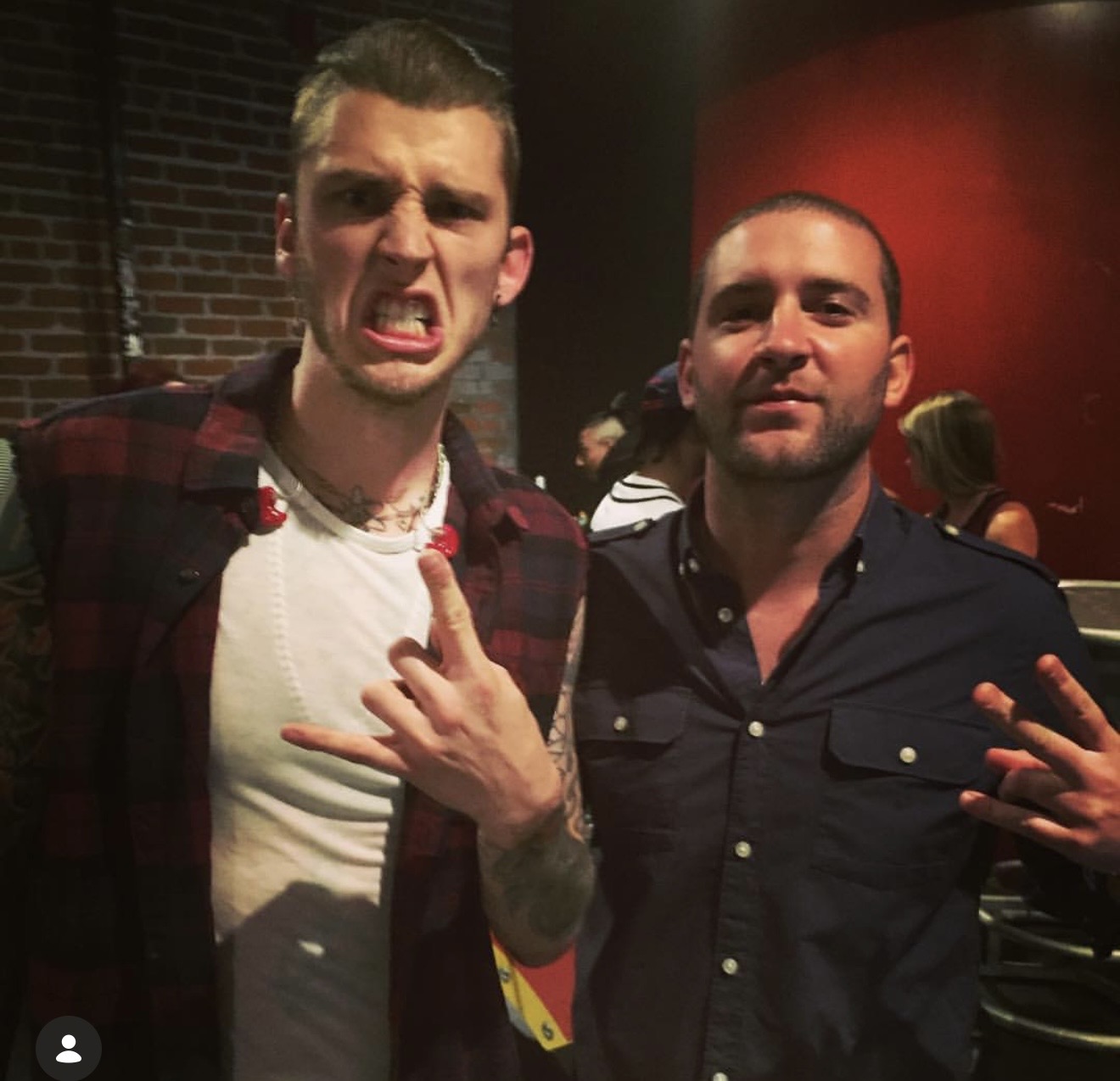 Two men posing indoors, one making a face and hand gesture, the other smiling with hand gesture, brick wall background.