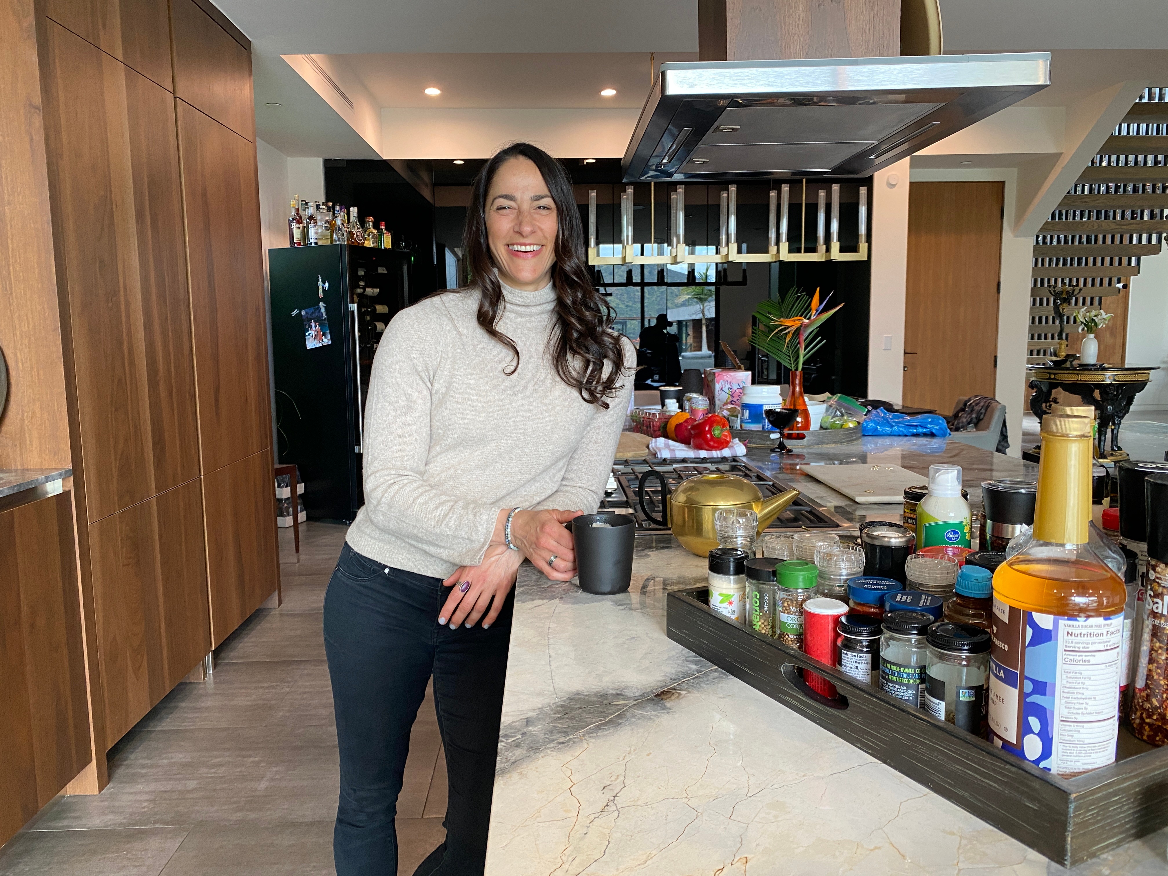 Woman with long dark hair smiling in a kitchen, wearing a white sweater and dark pants, standing near a marble countertop.
