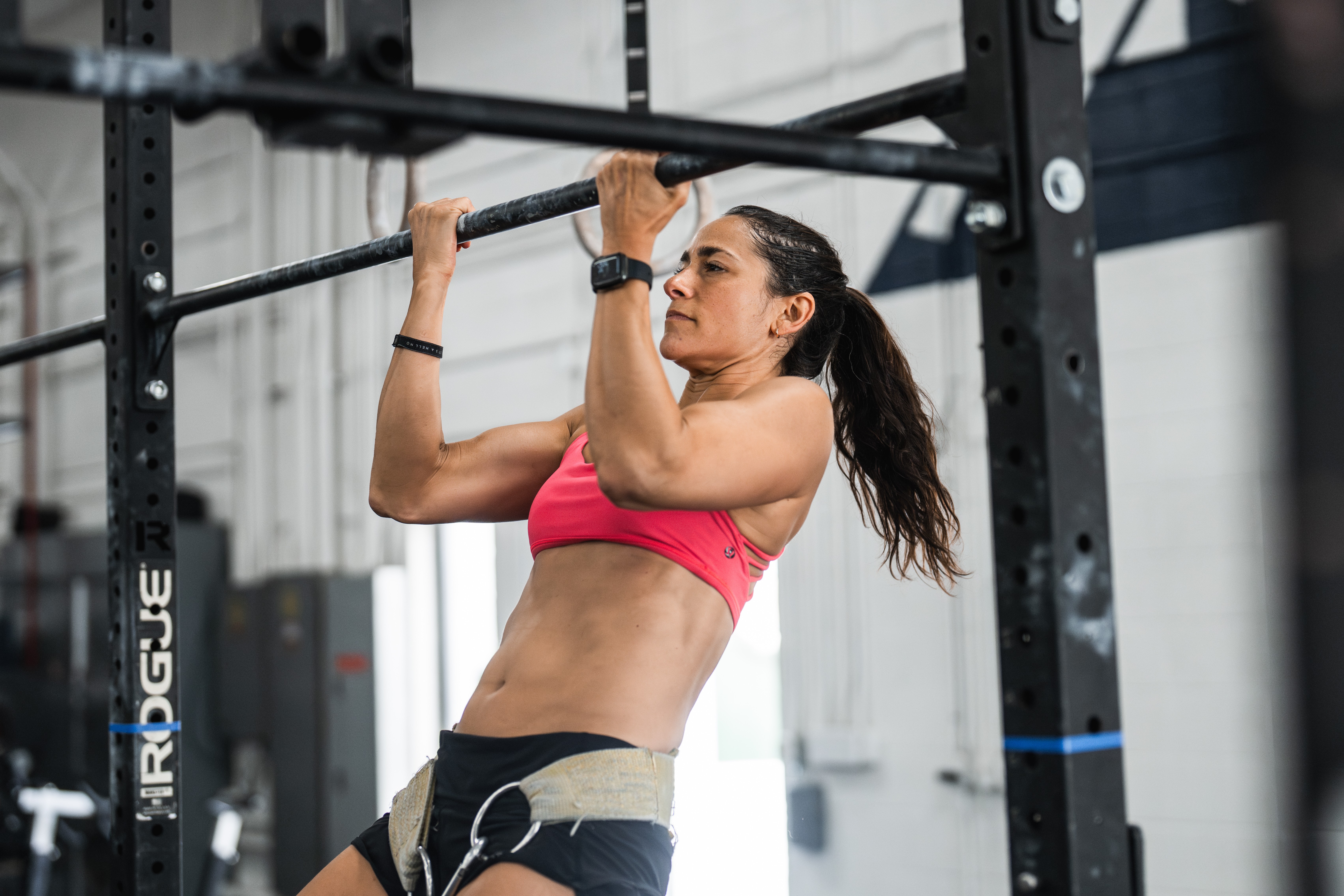Woman in pink sports bra and shorts doing pull-up exercise on gym equipment.