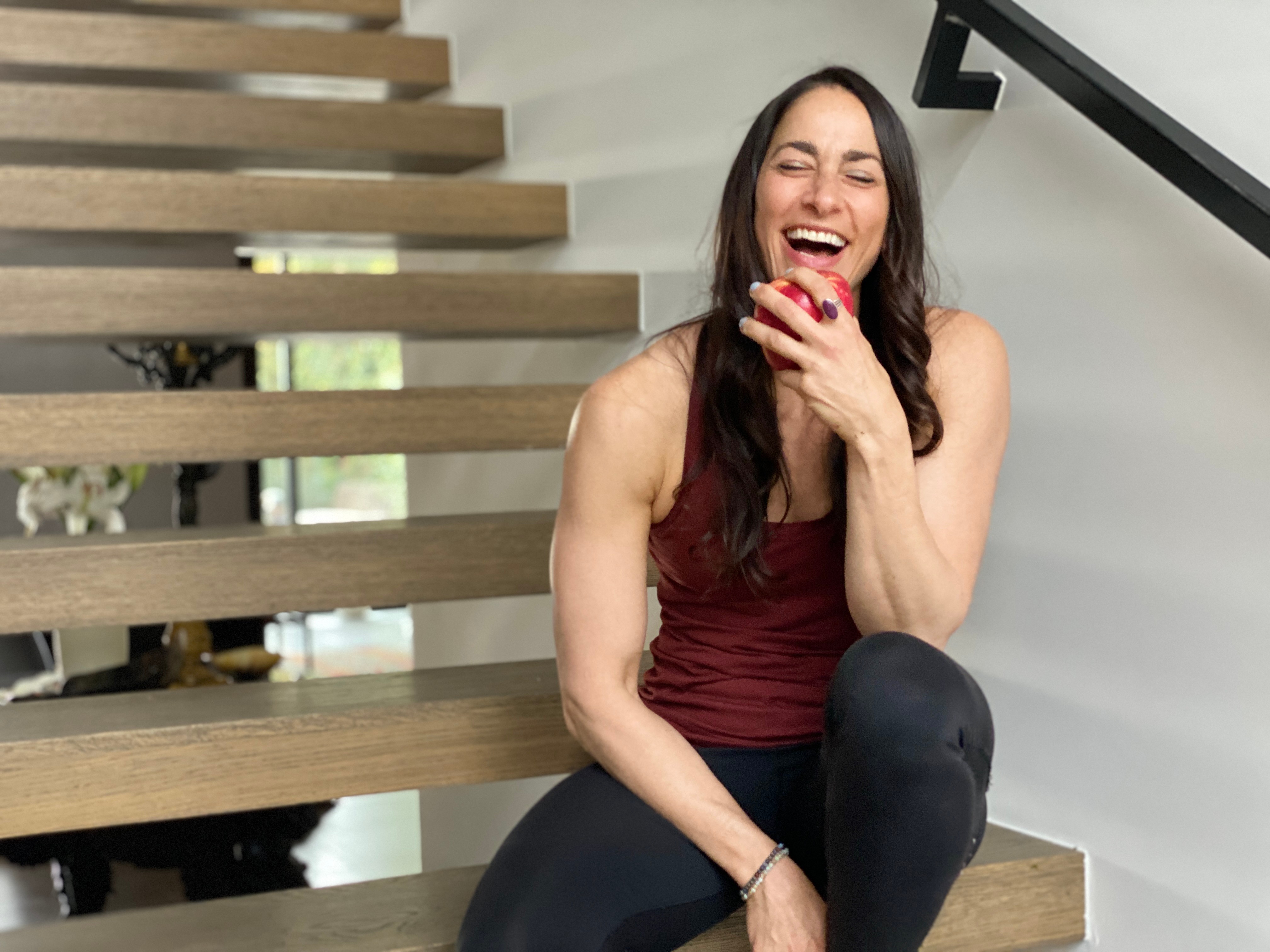 Woman sitting on stairs, smiling, holding an apple, wearing a sleeveless top and black pants.