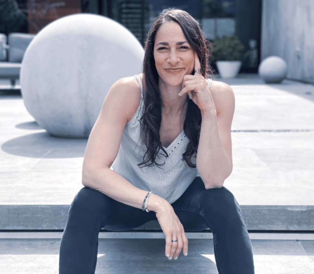Woman sitting outdoors on steps, smiling, with large spherical sculpture behind her, wearing a sleeveless top and dark pants.