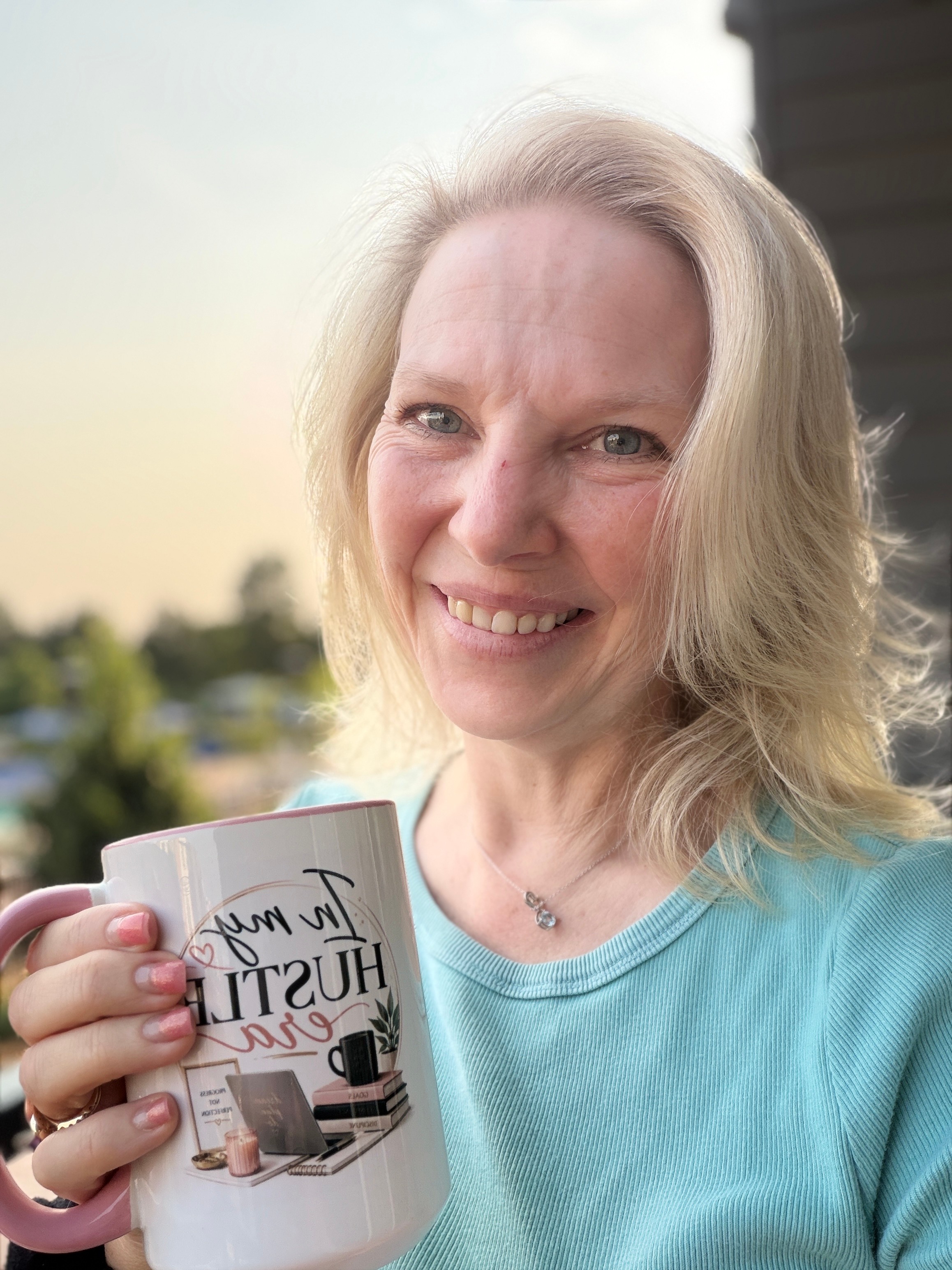 Smiling woman with blonde hair holding a mug outdoors, wearing a light blue shirt, with trees and sky in background.