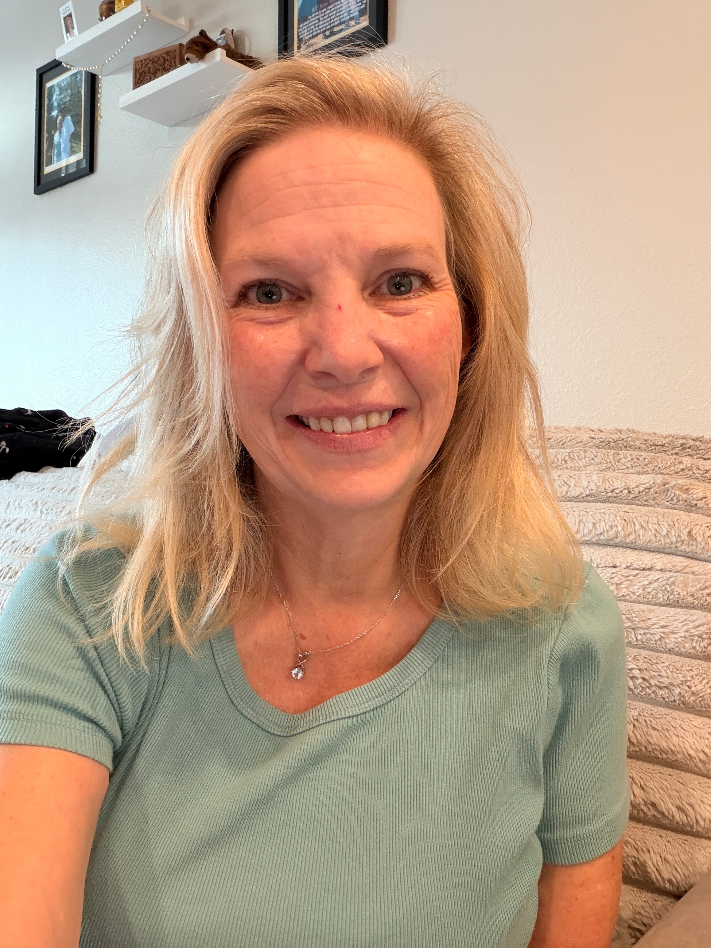 Woman with blonde hair smiling, wearing a light green shirt, sitting indoors with a beige cushion behind her.