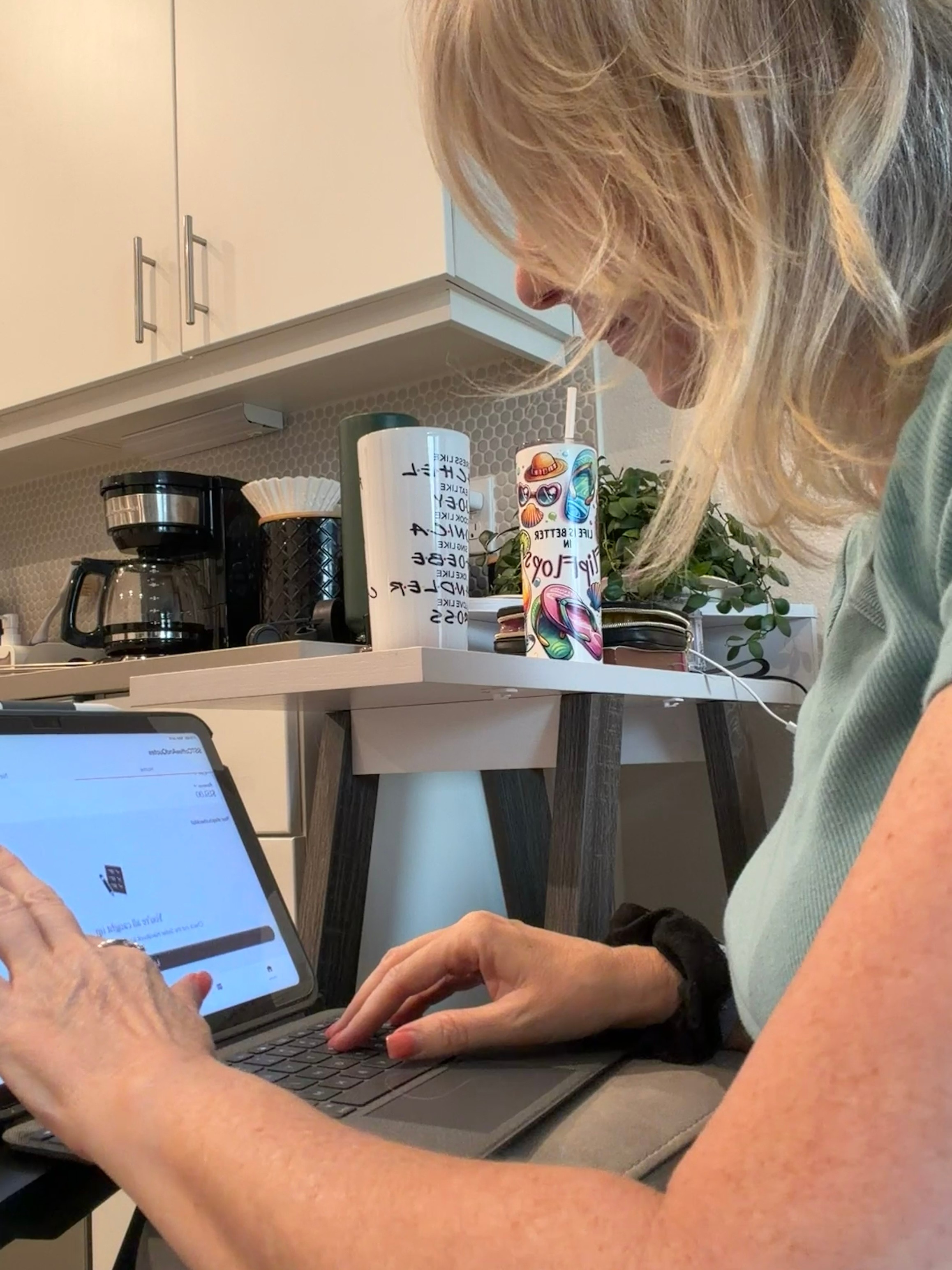 Woman with blonde hair using a laptop in a kitchen, with coffee maker and cups on the counter behind her.