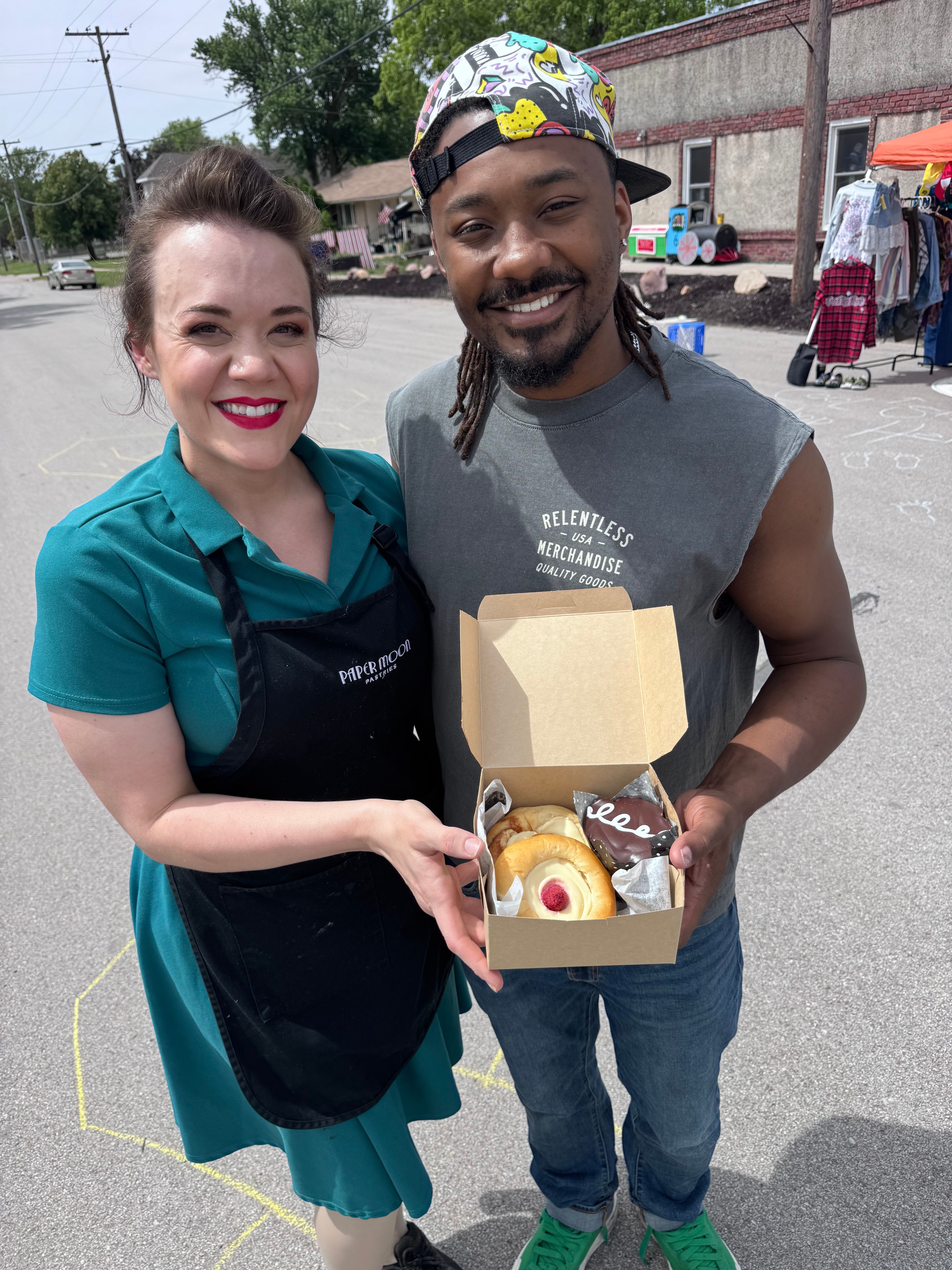 Two women standing outdoors, one holding a box with a cake and a drink, smiling at the camera.
