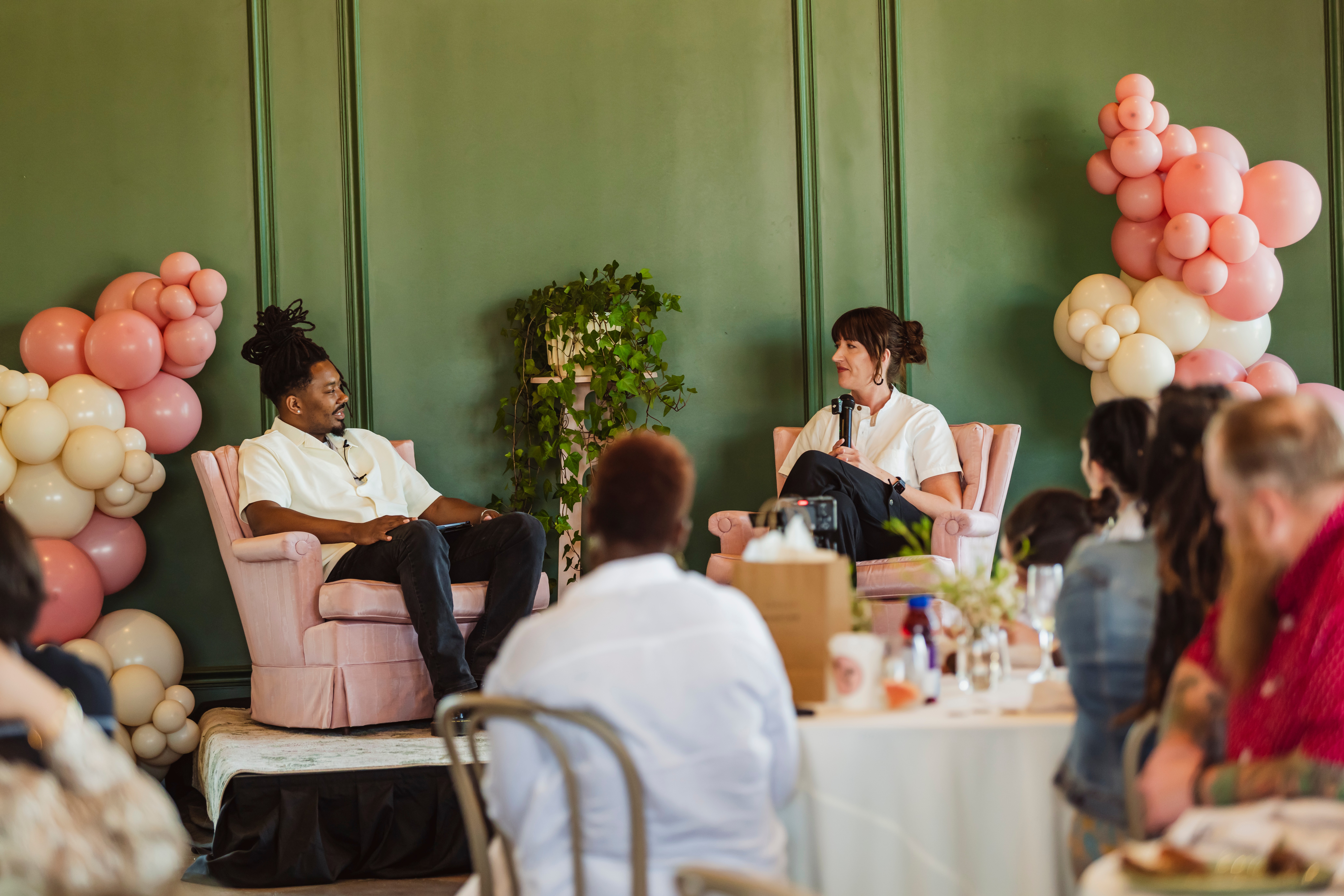 Two women sit on pink chairs in front of a green wall, engaging in conversation during a gathering with seated guests.