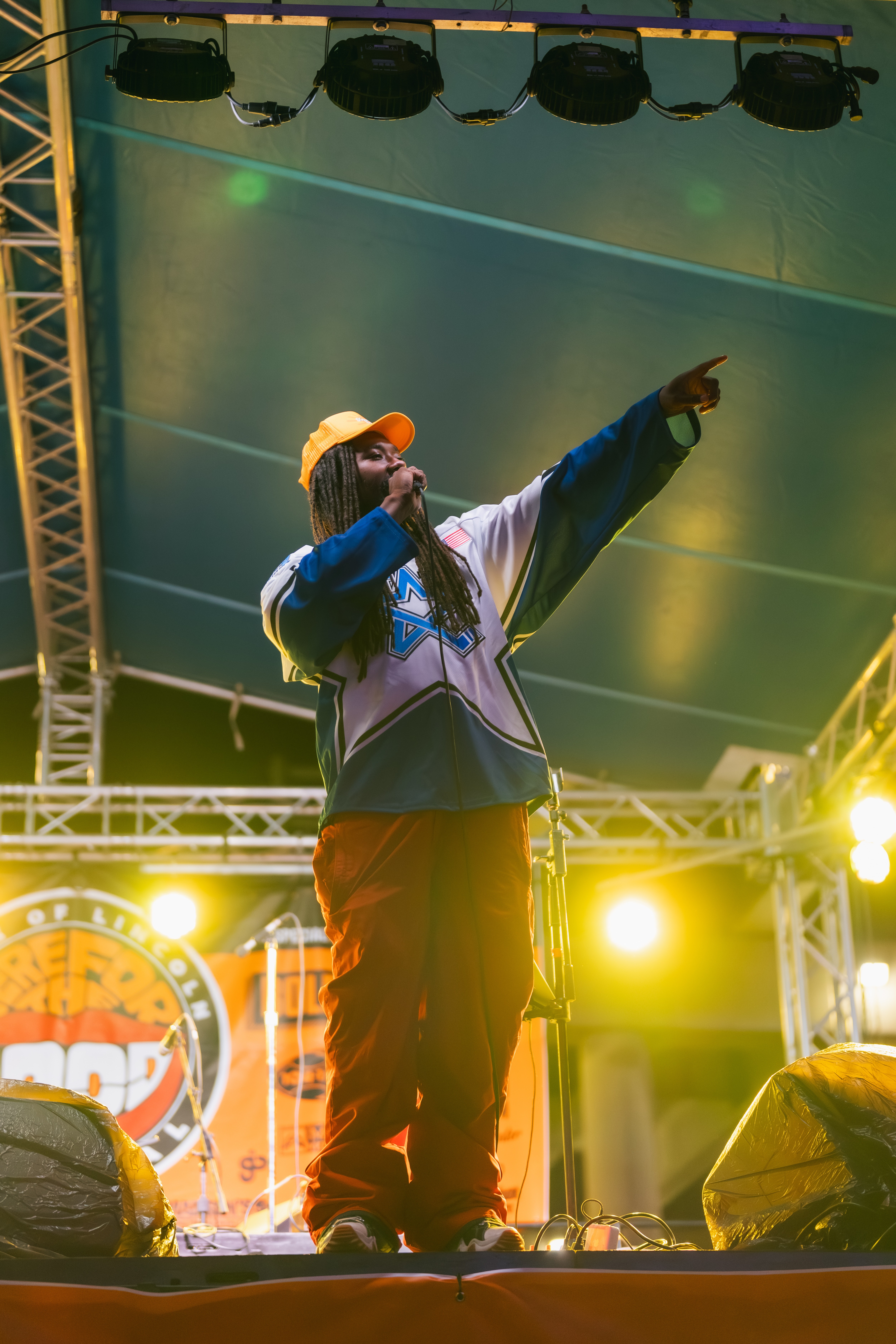 Performer on stage pointing upward, wearing a cap and jacket, with stage lights and equipment visible.