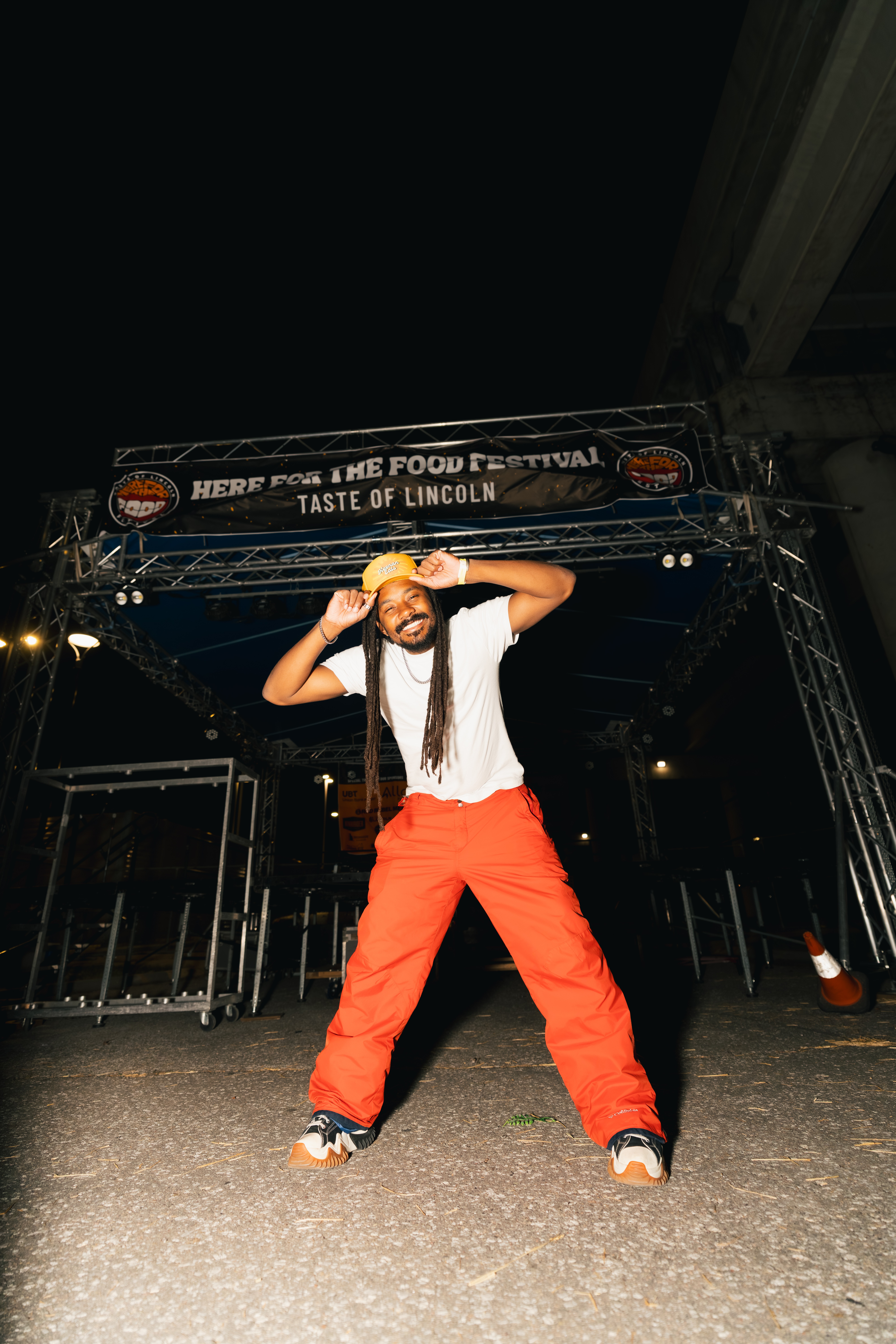 Person standing on stage at night, holding a hat, wearing a white shirt and red pants, with a festival banner overhead.