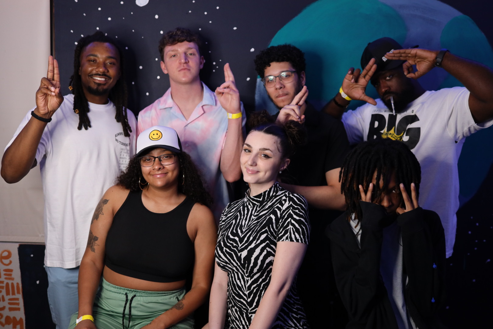 Group of seven diverse young people posing together, some making peace signs, in front of a space-themed background.