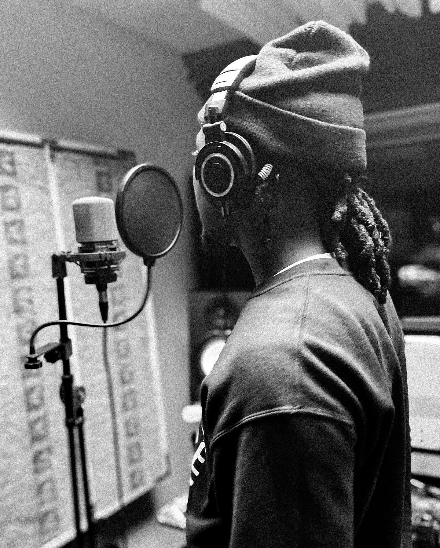 Person wearing a beanie and headphones in a recording studio, standing near a microphone, facing sideways.