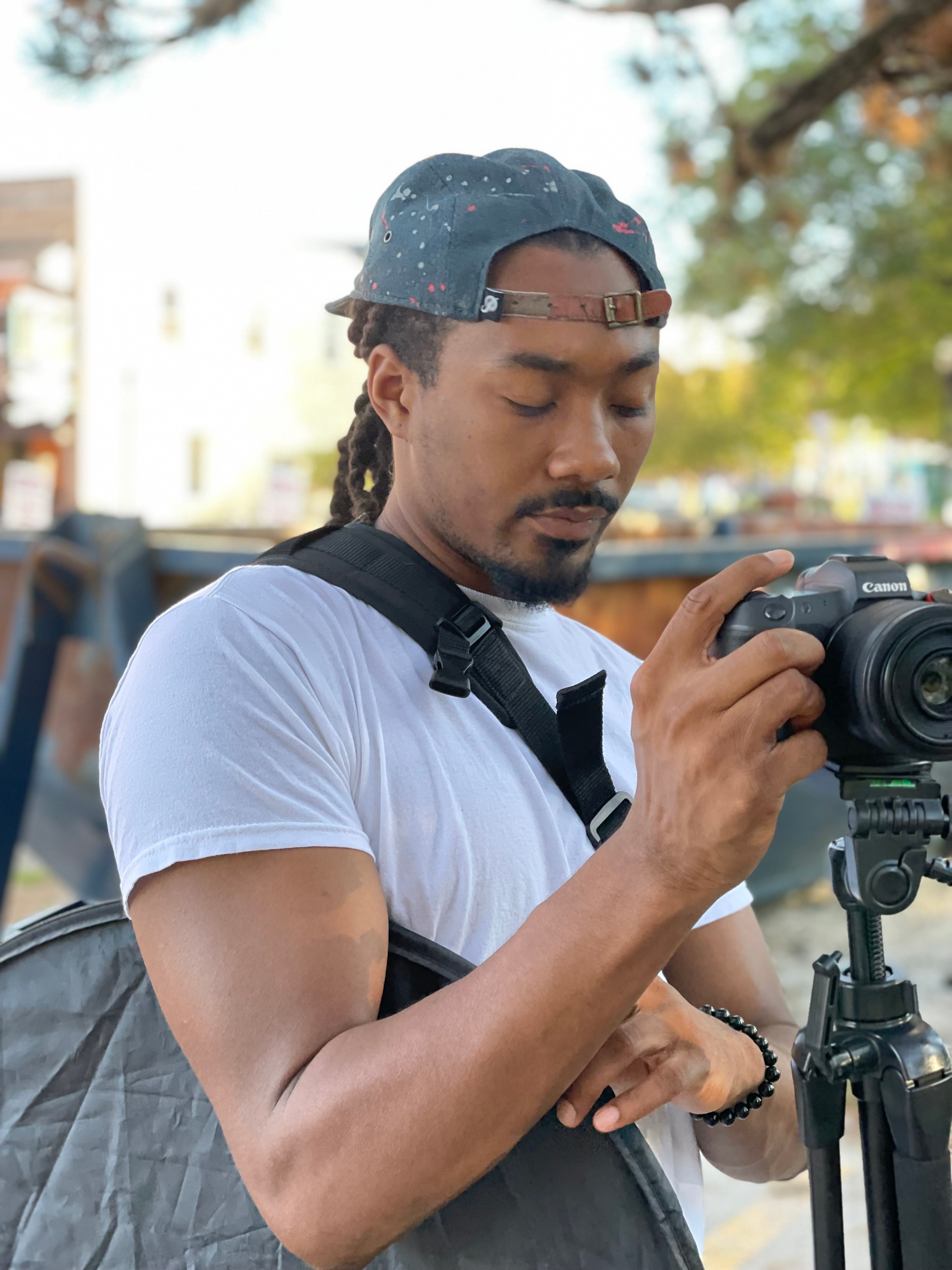 Man with a cap and camera outdoors, adjusting camera settings, trees and buildings in background.
