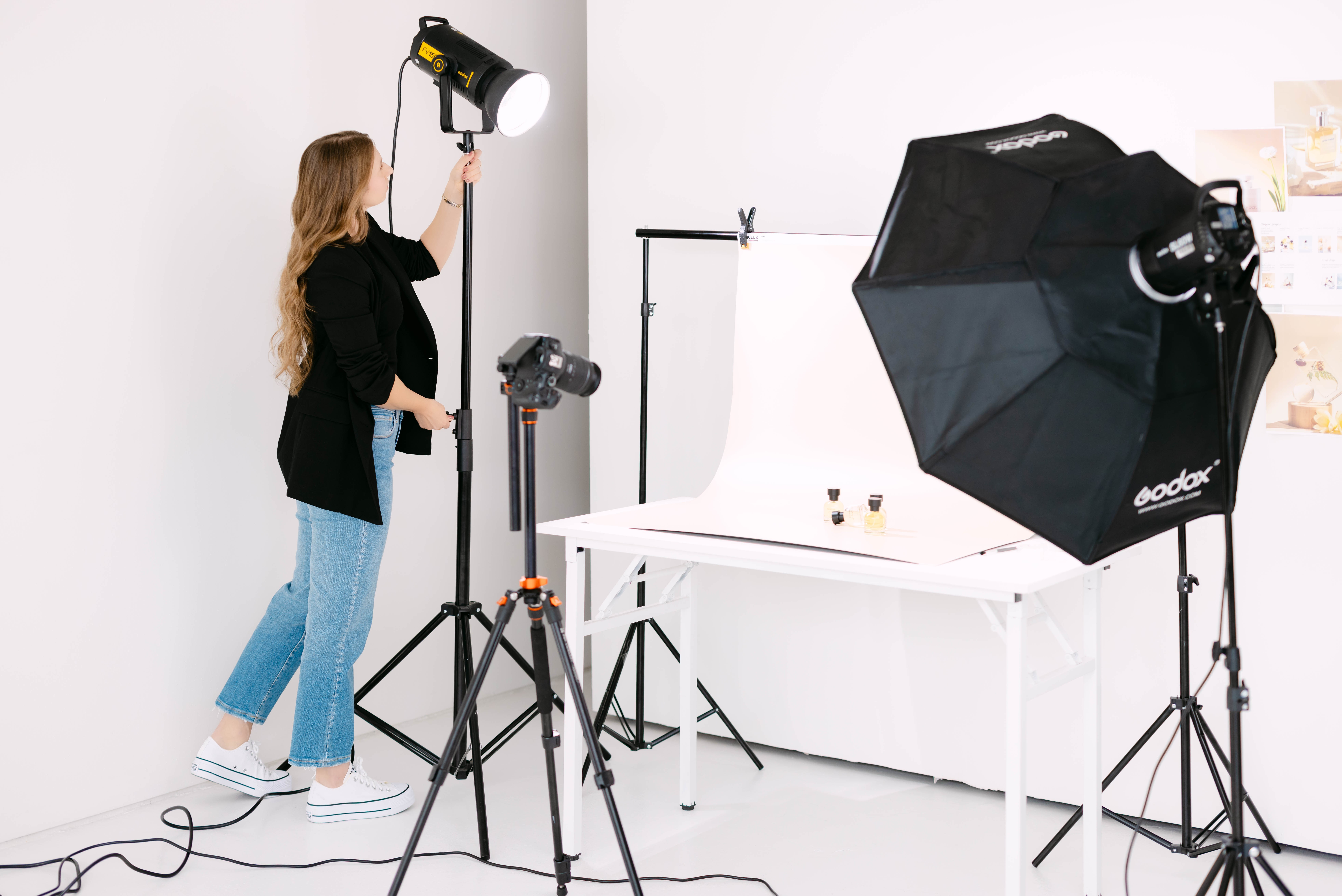 Woman adjusting studio lighting in photography setup with softbox and backdrop, white background, and equipment on stand.