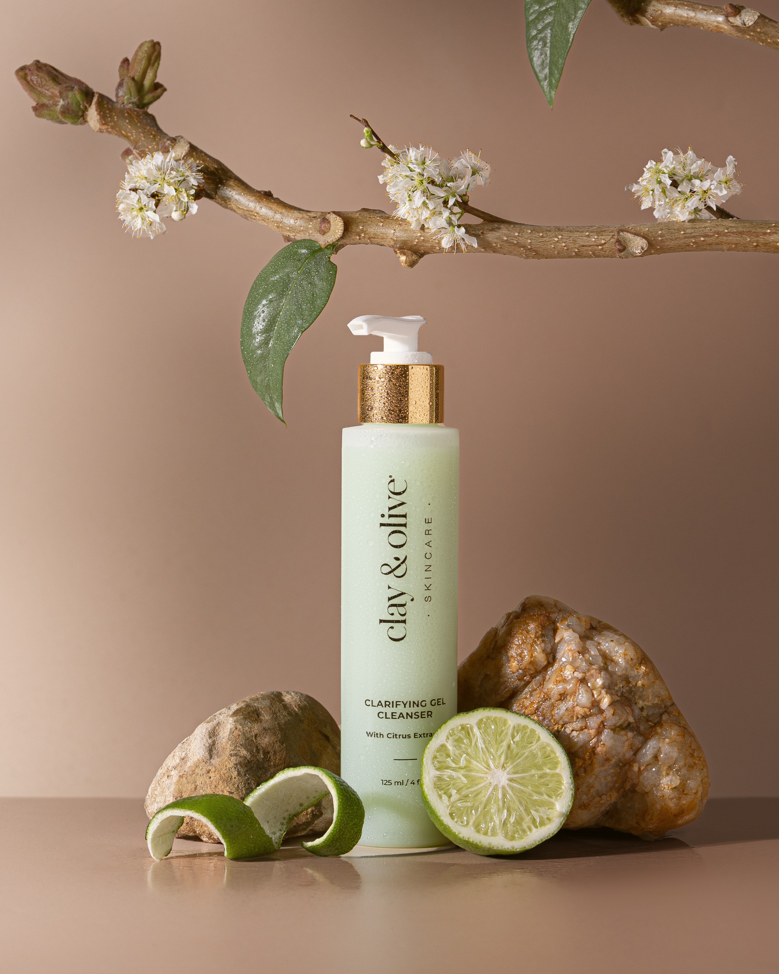 A bottle of clay & olive cleansing foam with lime slices, rocks, and a flowering branch against a neutral background.