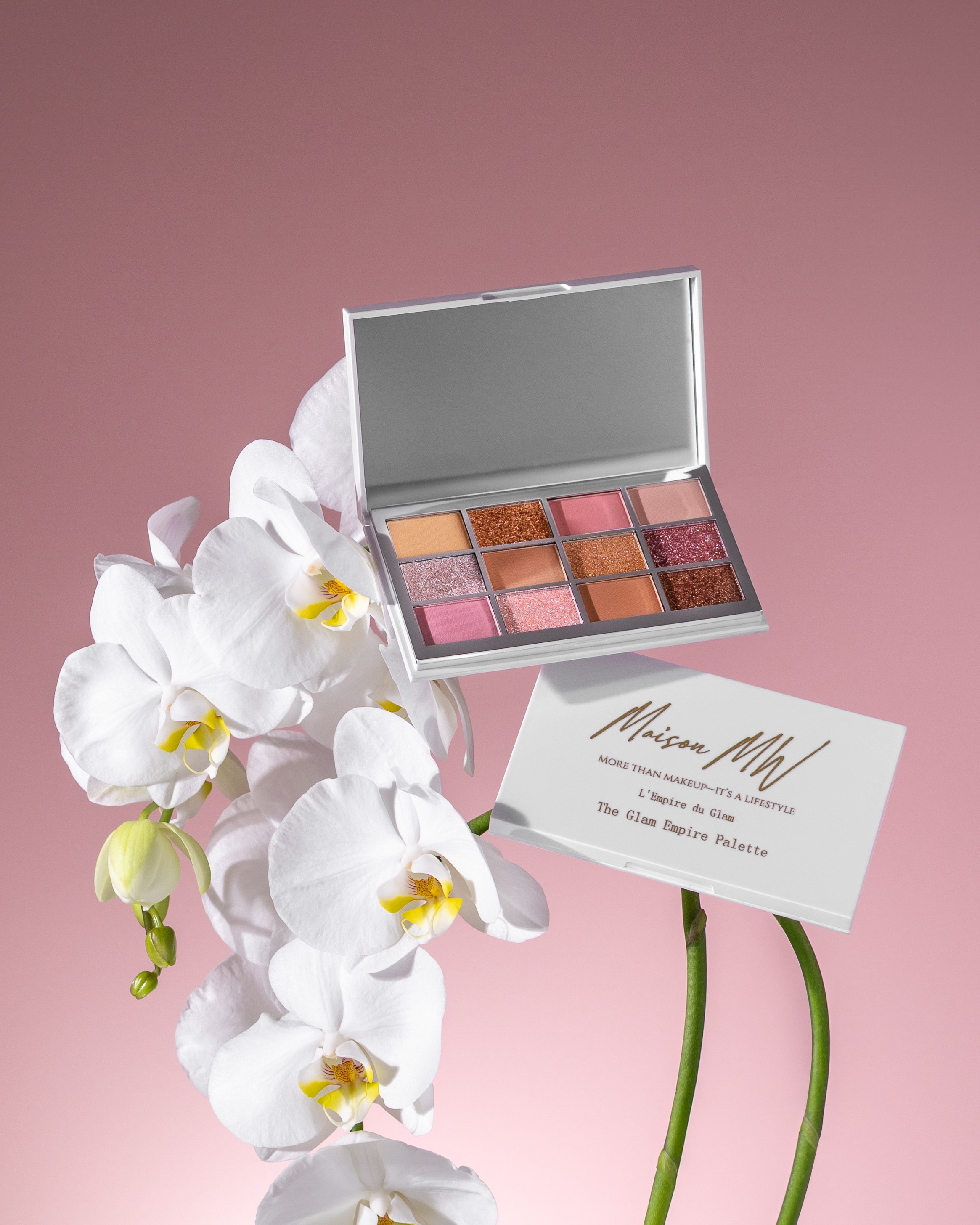Open eyeshadow palette with nine shades, white orchids, and a business card on a pink background.