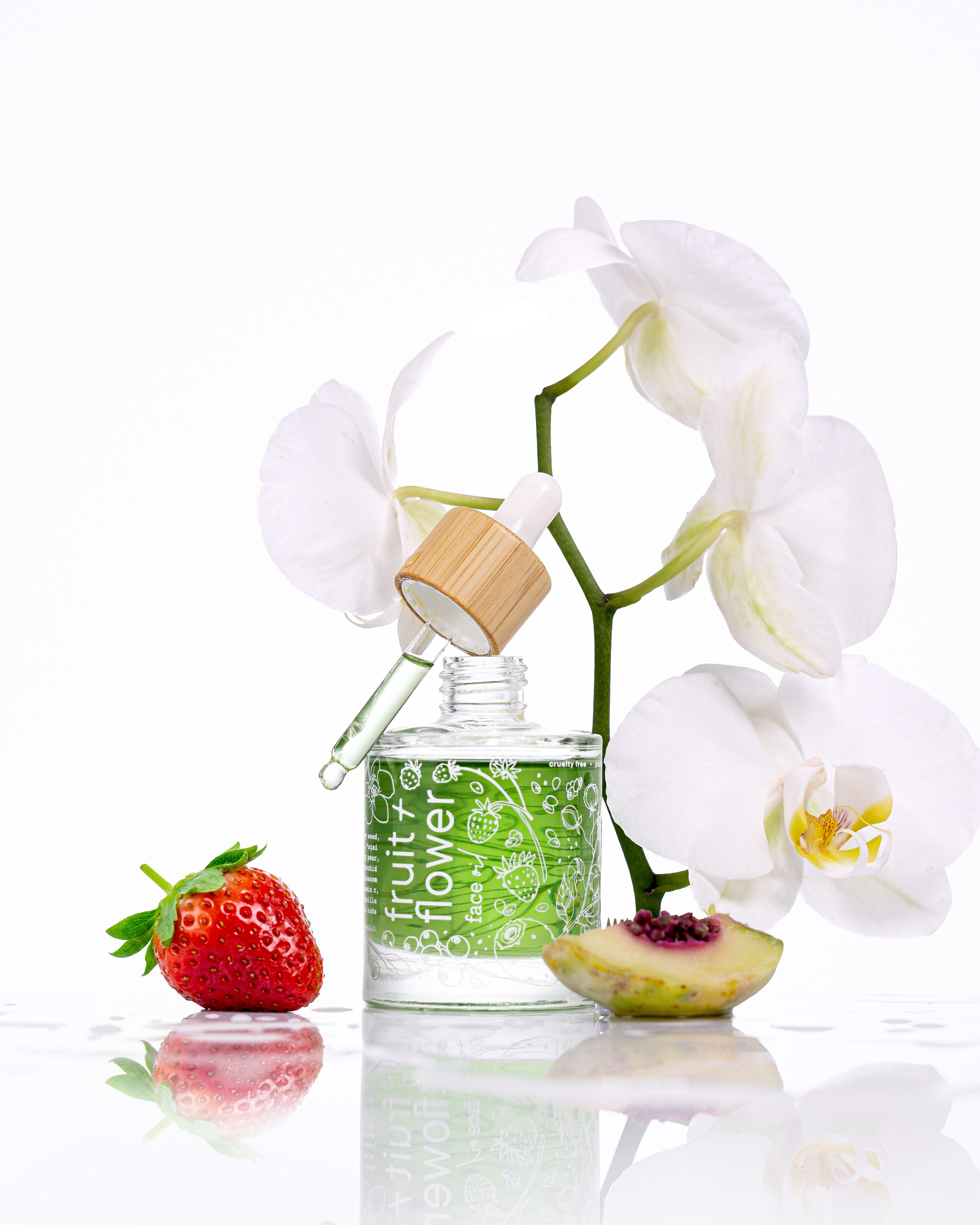 White orchid flower in a green bottle, strawberry, and a sliced fruit on a reflective surface.